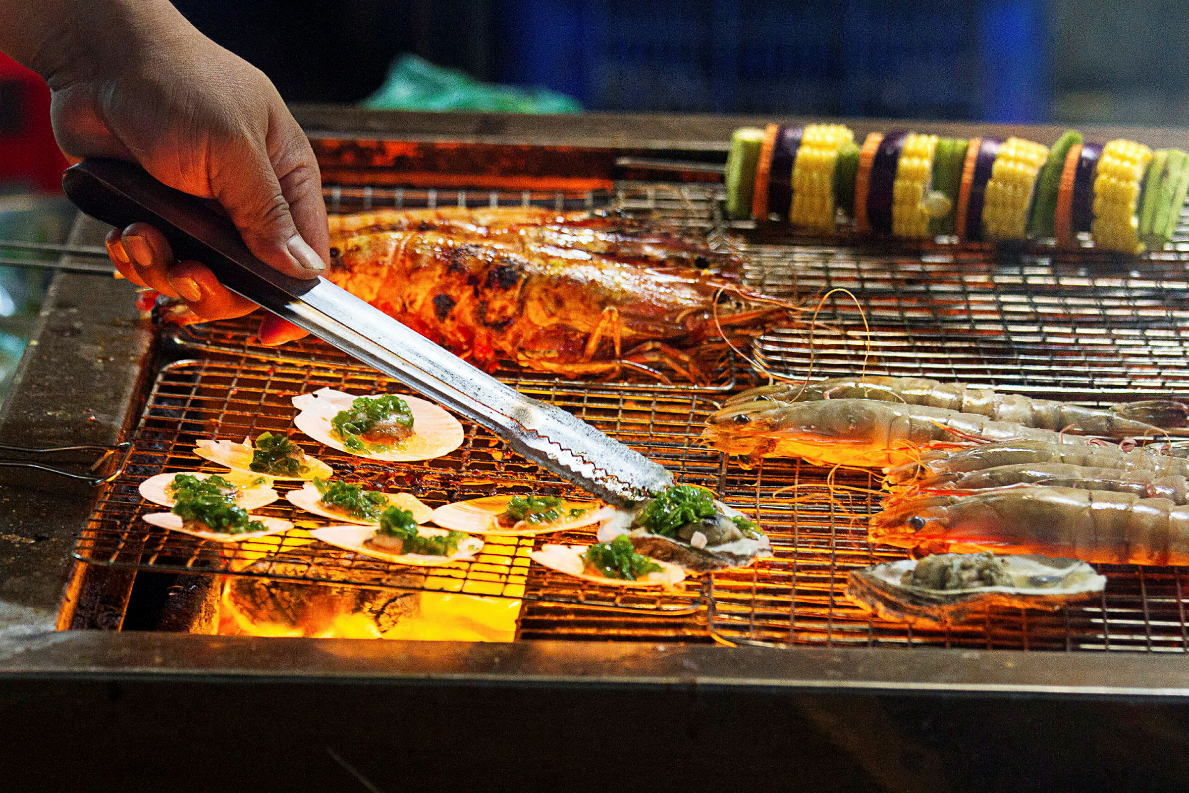 En grill med skaldyr og grøntsagsspyd, hvor en hånd håndterer maden med en tang. (AI-genereret tekst)