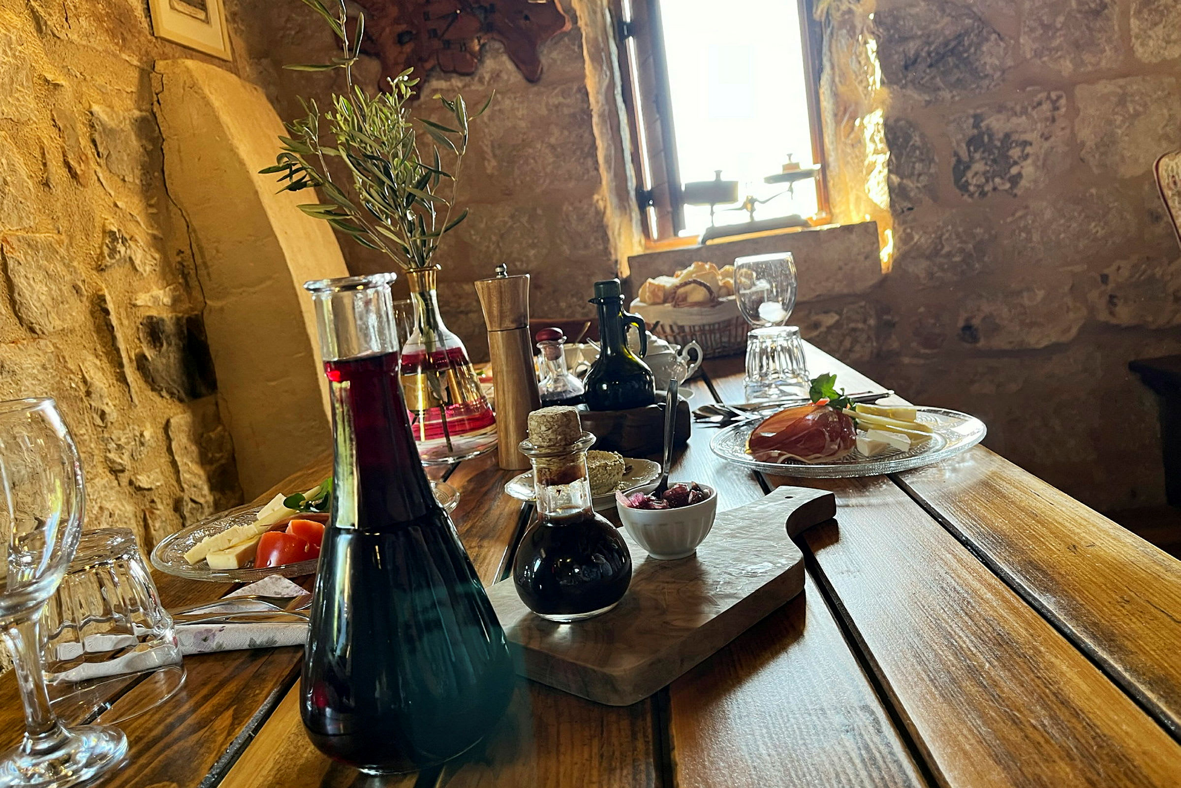 Et rustikt træbord med forskellige serveringer, herunder olivenolie, vin, brød og pålæg, samt dekorationer i et ældre miljø. (AI-genereret tekst)