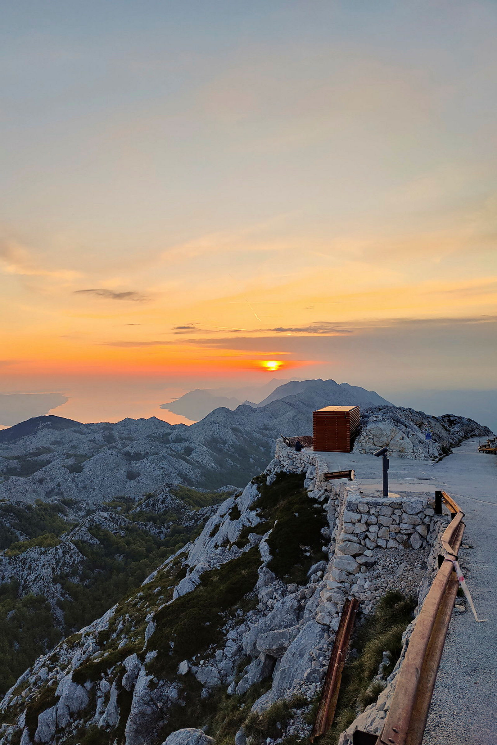 Billede af en bjergtop under solnedgang med stenmur, en gangsti og et rækværk i forgrunden. (AI-genereret tekst)