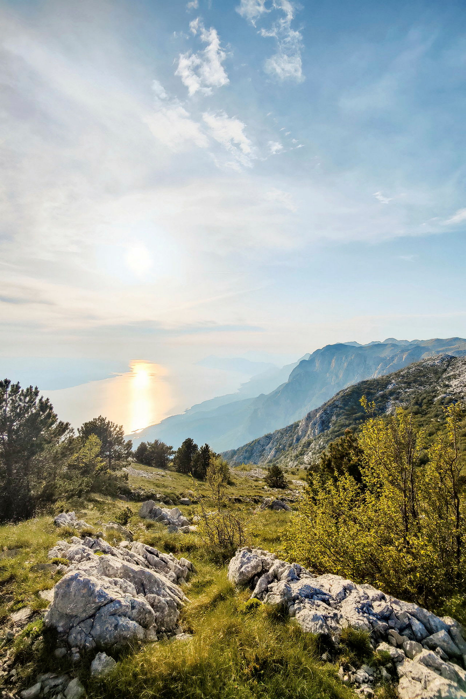 Udsigt over et bjerglandskab med vegetation, klipper og træer, og havet i baggrunden ved solnedgang. (AI-genereret tekst)