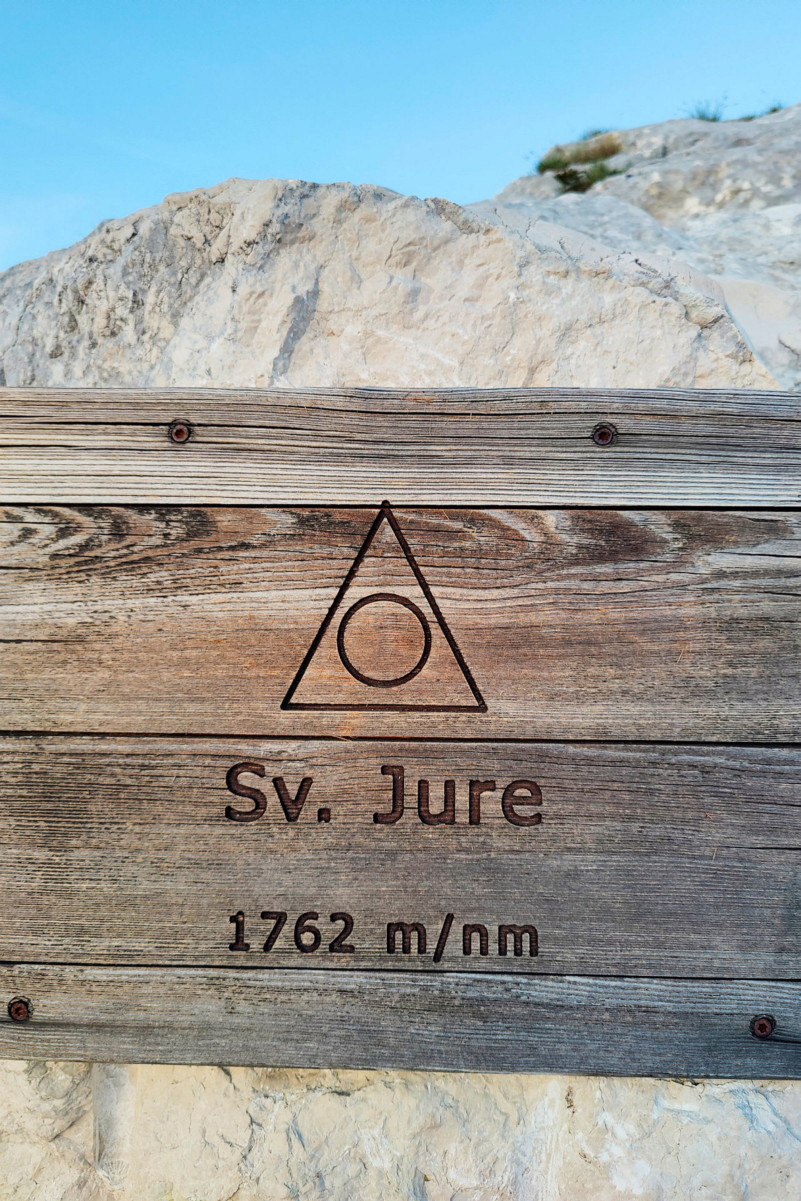 Træskilt med teksten 'Sv. Jure 1762 m/nm' mod en baggrund af klipper og blå himmel. (AI-genereret tekst)