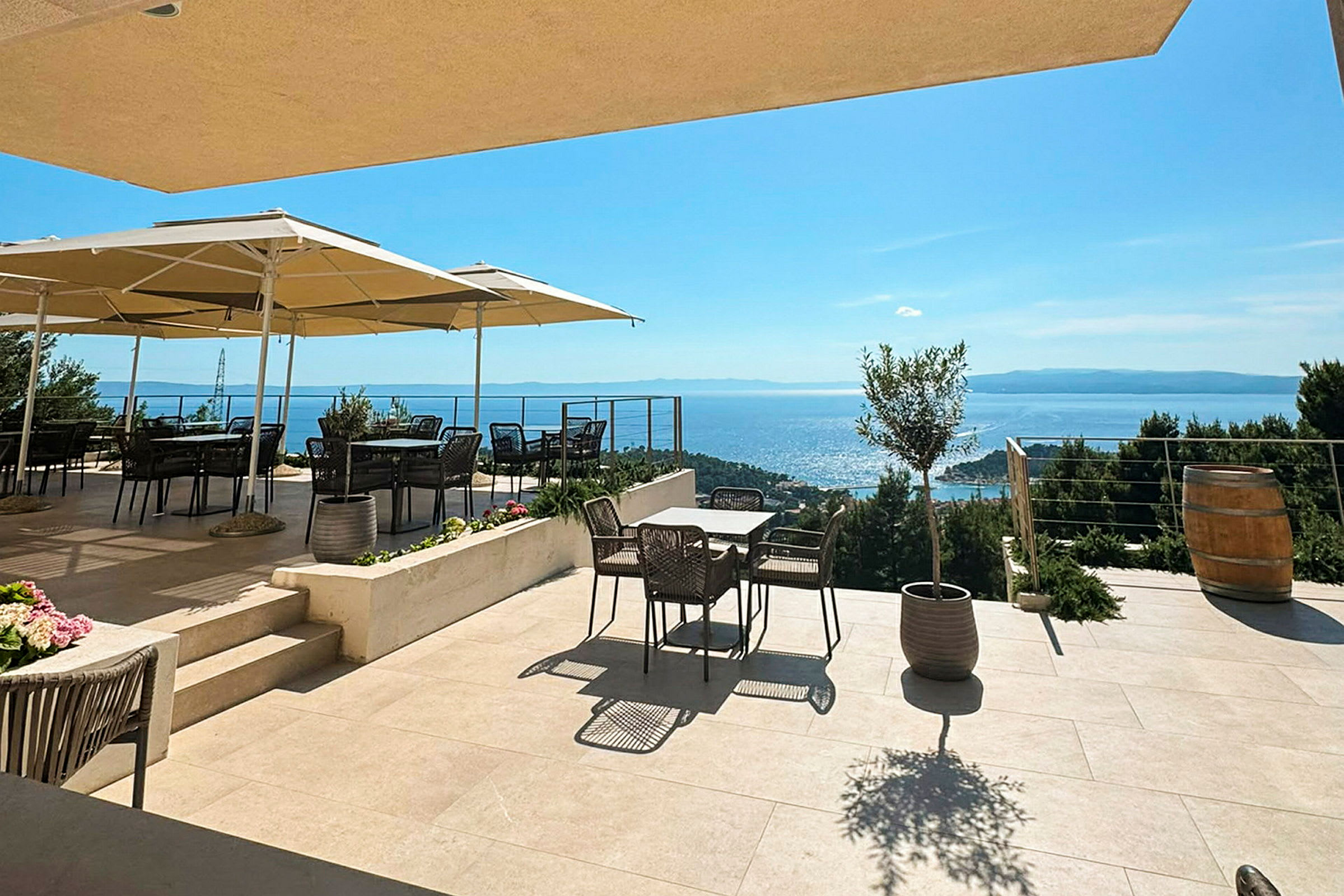 En terrasse med borde, stole og parasoller, med udsigt over havet og det omkringliggende landskab. (AI-genereret tekst)
