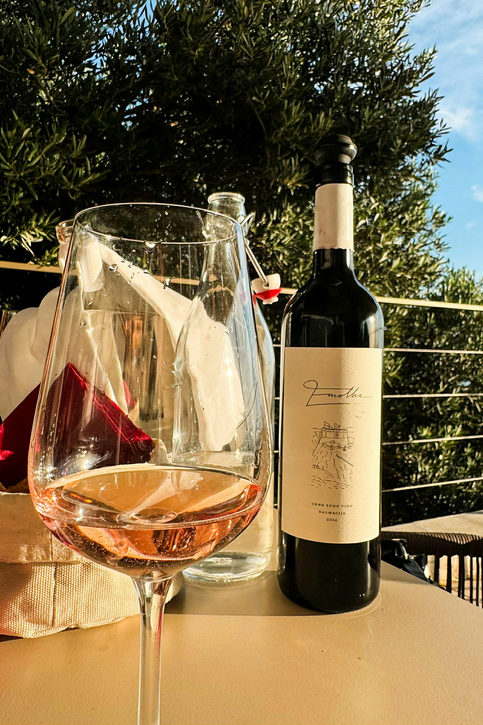 Et glas rosévin og en vinflaske står på et bord i en udendørs indstilling med grønne blade i baggrunden. (AI-genereret tekst)