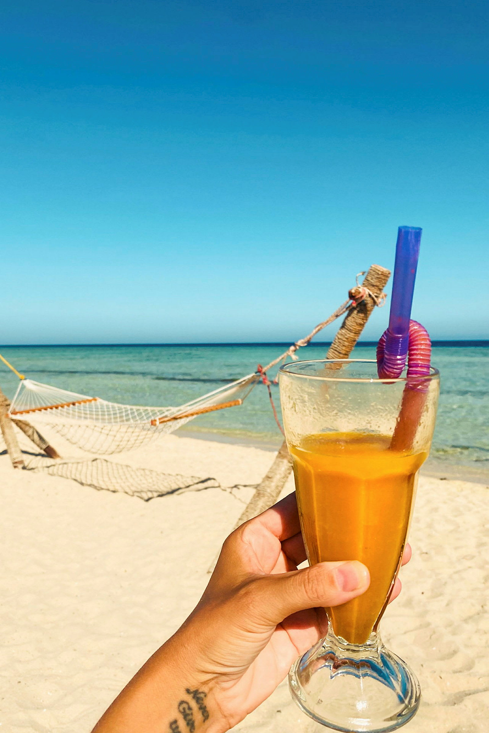 Et glas med gul drik holdes af en hånd på en strand med en hængekøje og hav i baggrunden. (AI-genereret tekst)