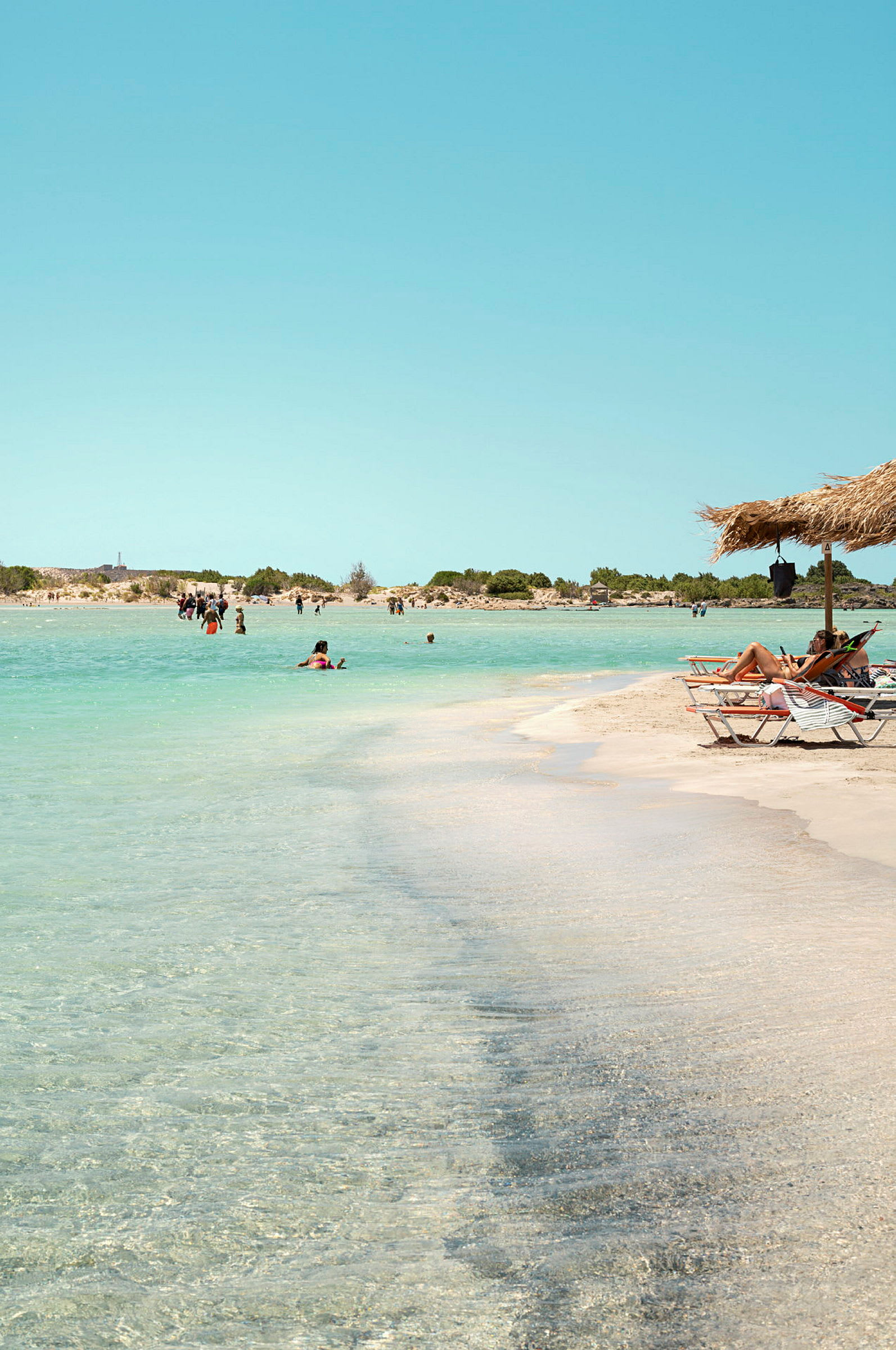 En strand med hvid sand og klart vand, omgivet af lave buske. Flere personer bader i havet, og nogle sidder i solsenge under en parasol. (AI-genereret tekst)