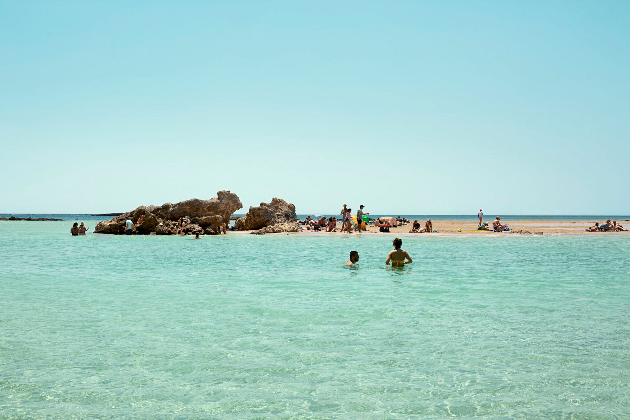 En strand med klipper og krystalklart vand. Mennesker nyder stranden og bader i havet. (AI-genereret tekst)