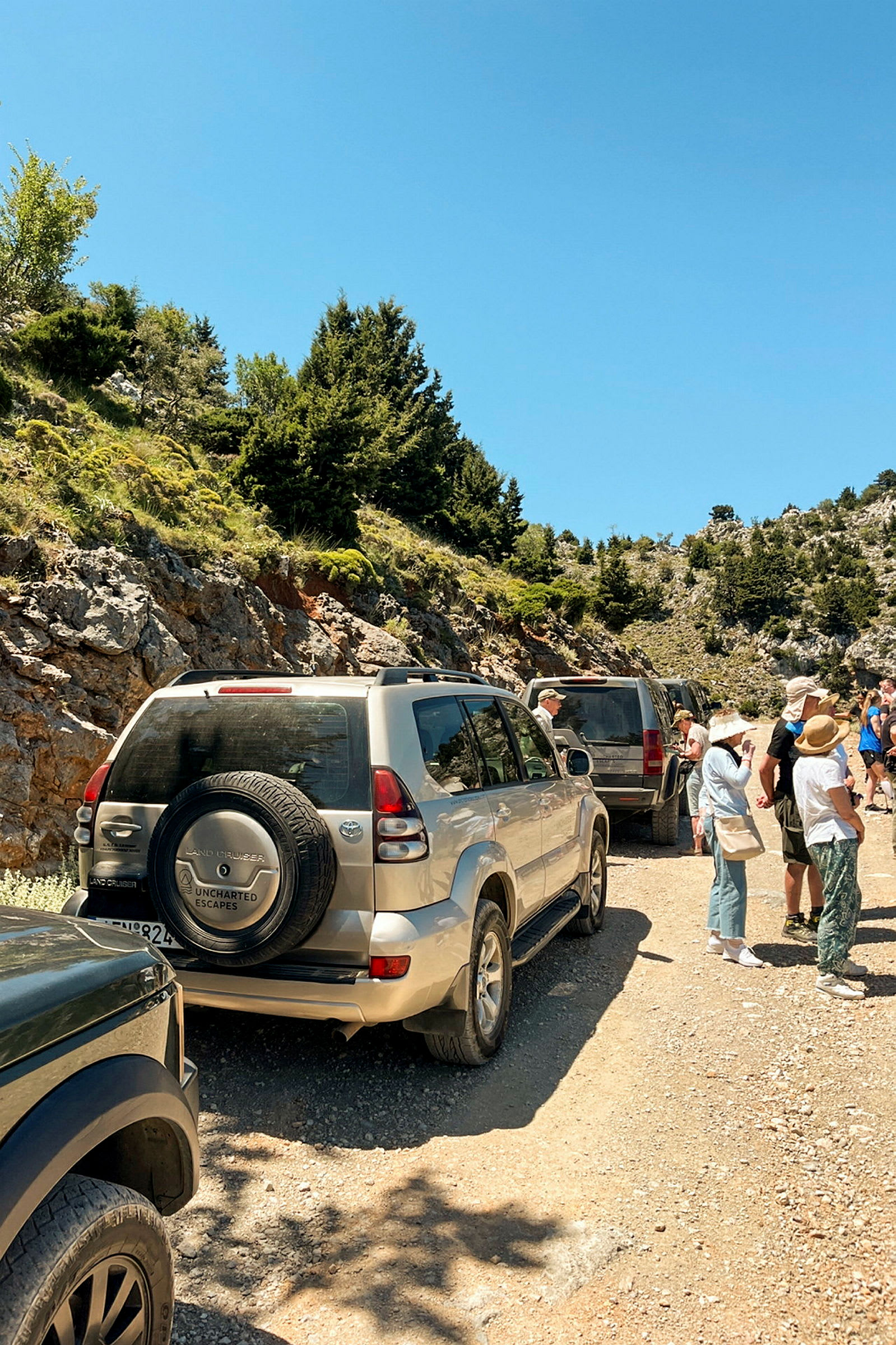 En gruppe terrængbiler parkeret ved en grusvej omgivet af bjerge og grønt landskab, hvor folk samles ved siden af vejen. (AI-genereret tekst)