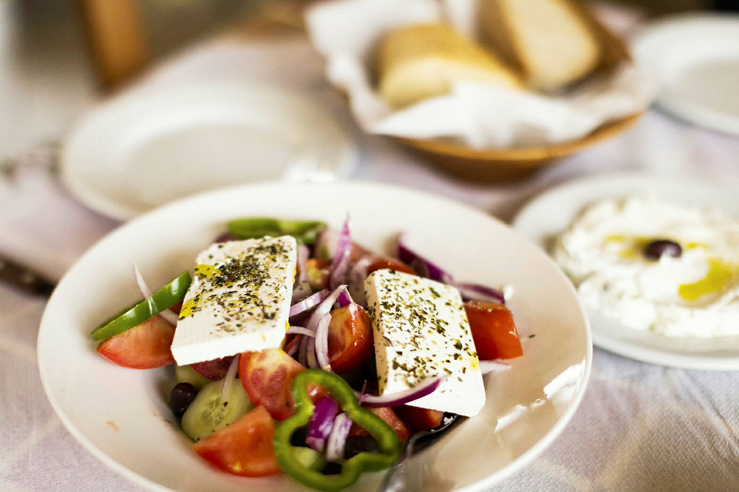 En tallerken med græsk salat indeholdende tomater, agurk, peberfrugt, løg og fetaost. I baggrunden ses brød og en tallerken med yoghurt og olivenolie. (AI-genereret tekst)
