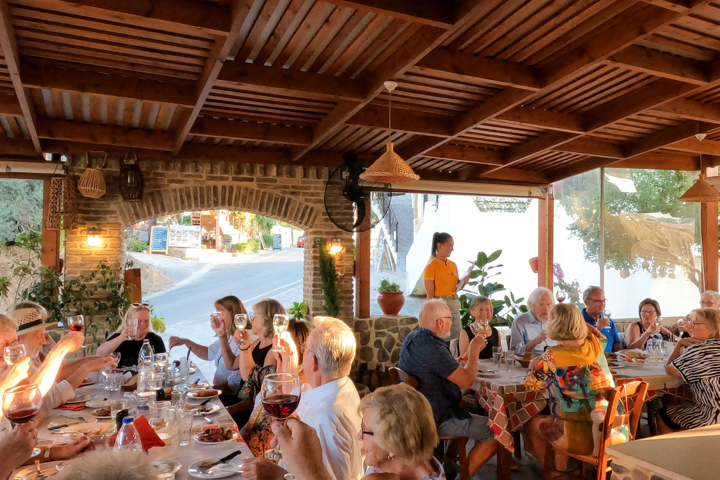 En gruppe mennesker sidder omkring et langbord på en udendørs restaurant under en træpergola. (AI-genereret tekst)