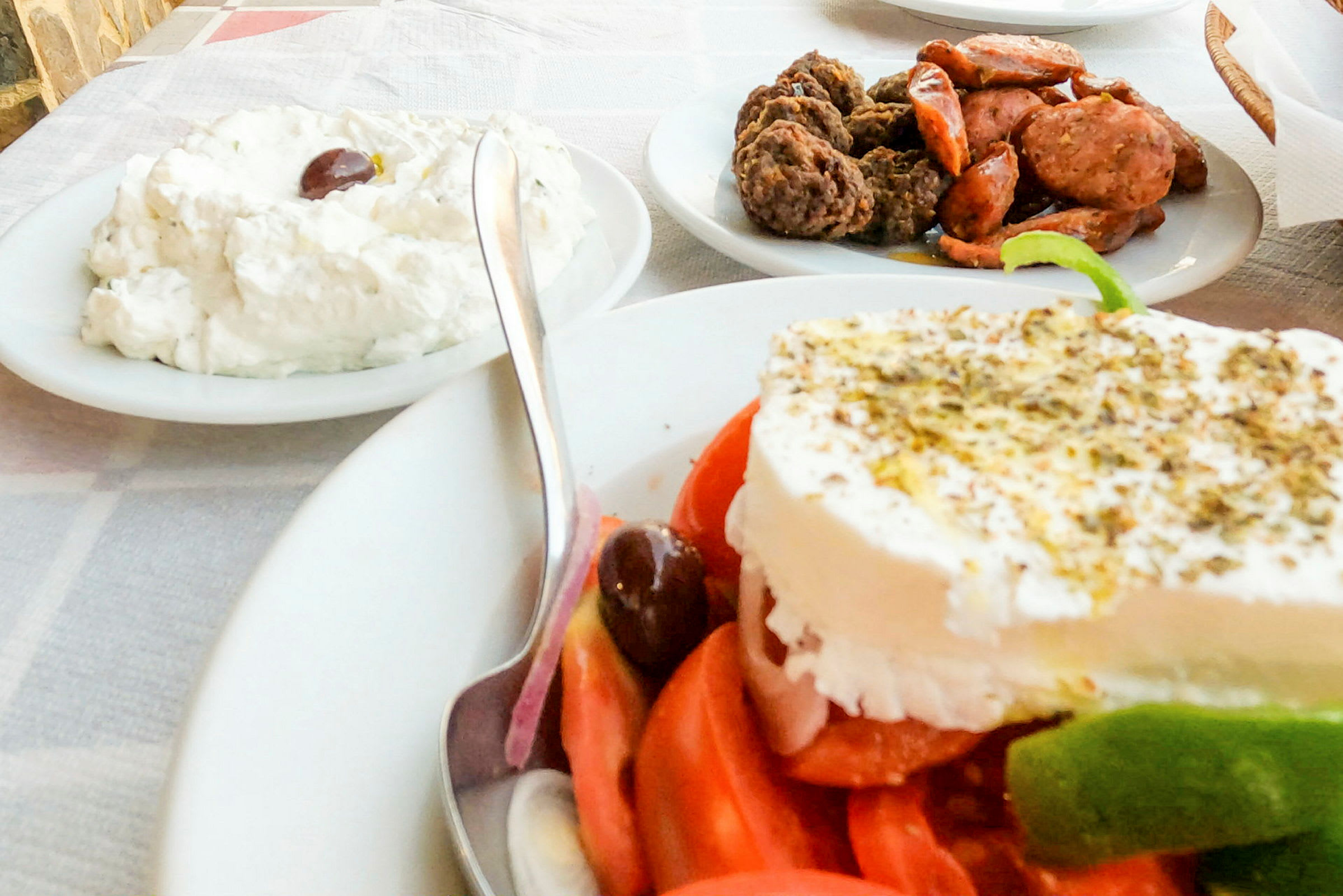 En tallerken med en græsk salat toppet med fetaost, en skål med tzatziki og en tallerken med kød og pølse på et bord. (AI-genereret tekst)