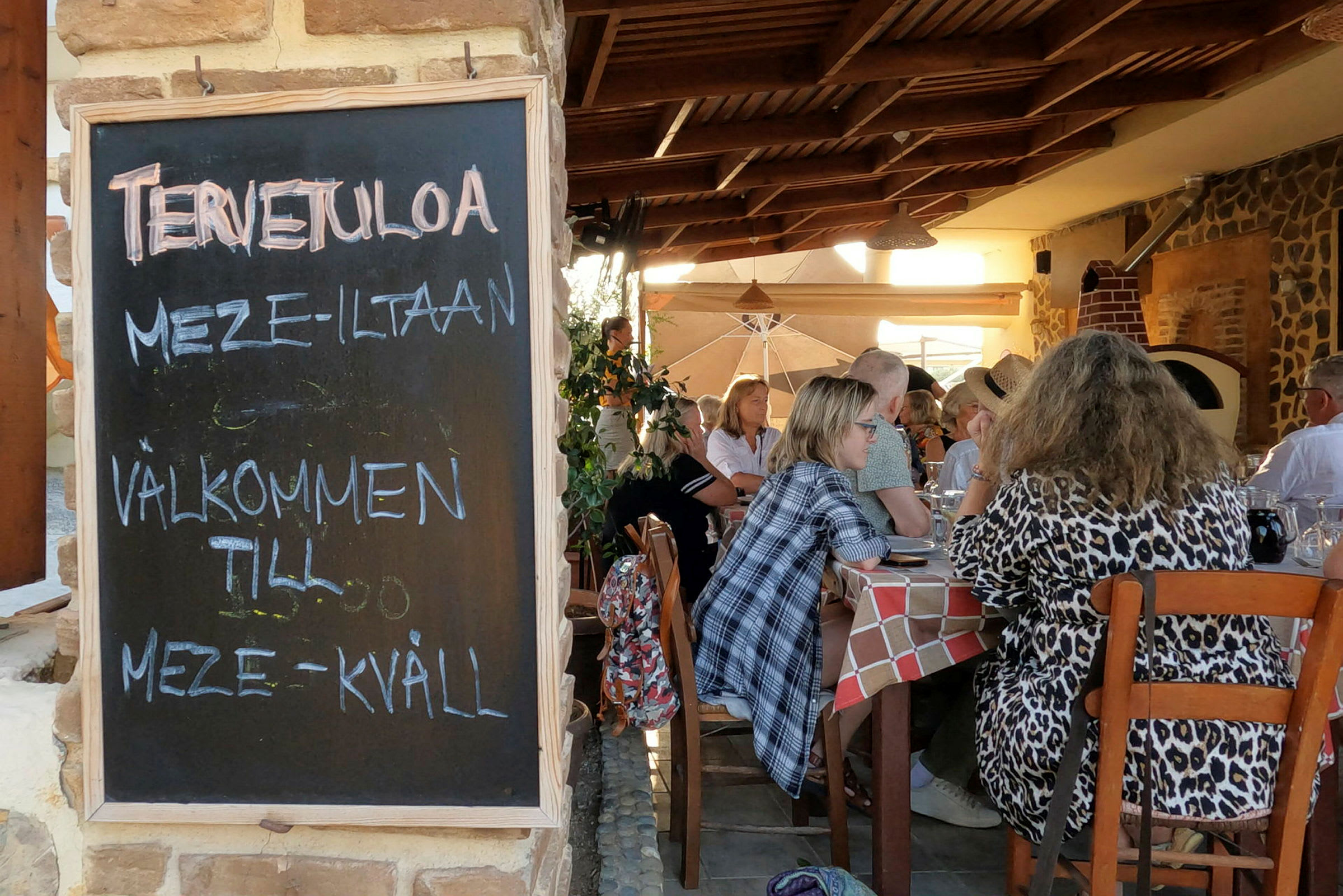 En sort tavle med teksten "Velkommen til meze-aften" står ved indgangen til en restaurant med gæster siddende ved borde. (AI-genereret tekst)