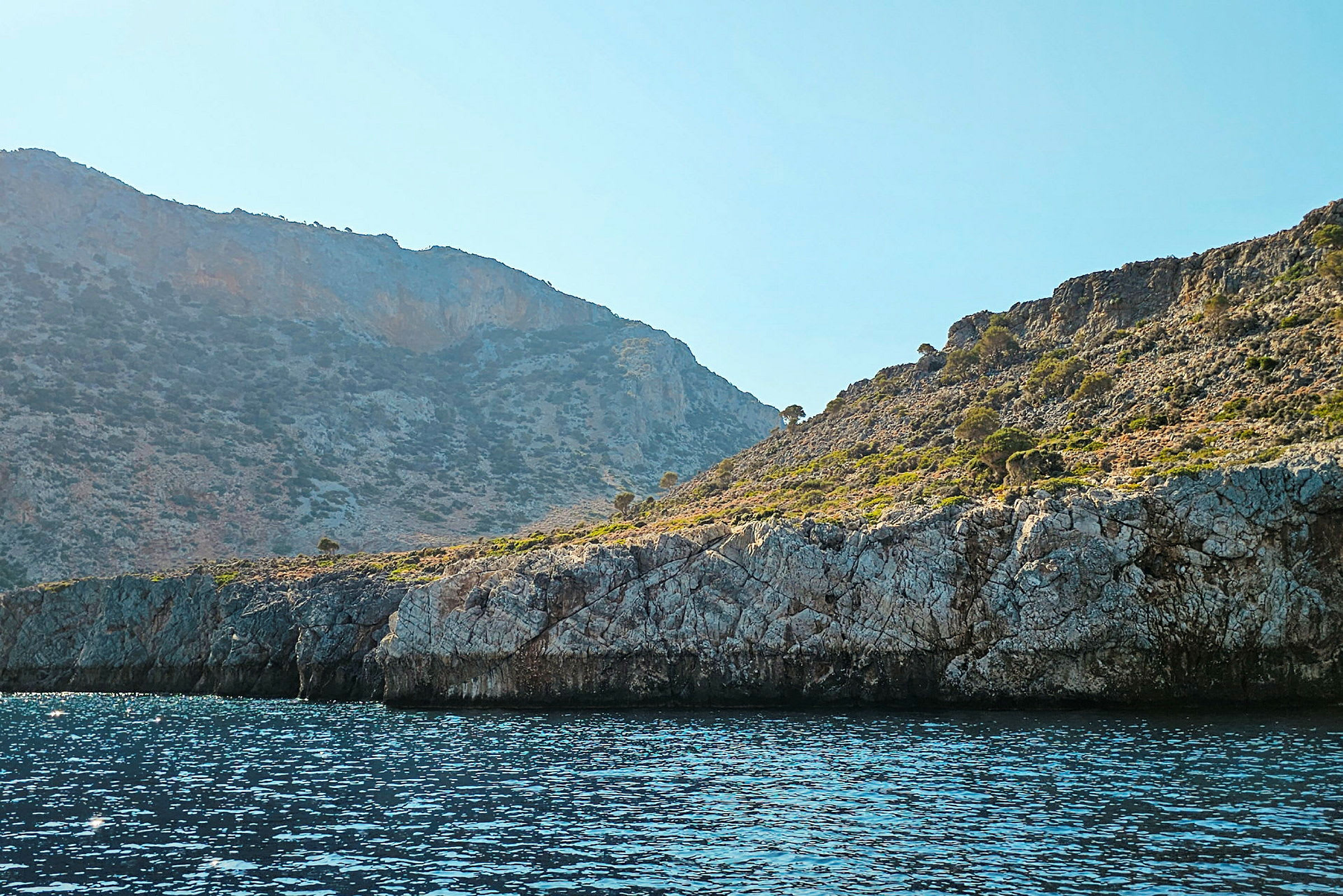 En klippefyldt kystlinje med havet i forgrunden og bjerge med vegetation i baggrunden. (AI-genereret tekst)
