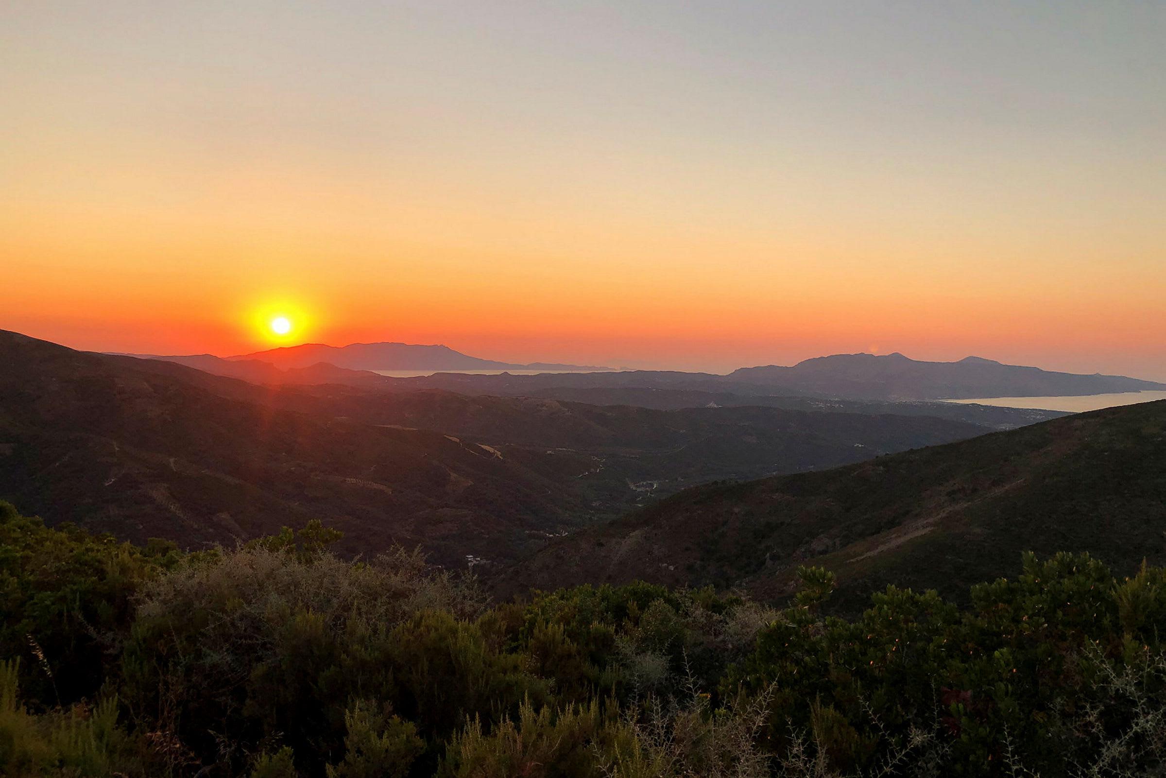 En solnedgang over et bjergrigt landskab med lav vegetation og havet i baggrunden. (AI-genereret tekst)