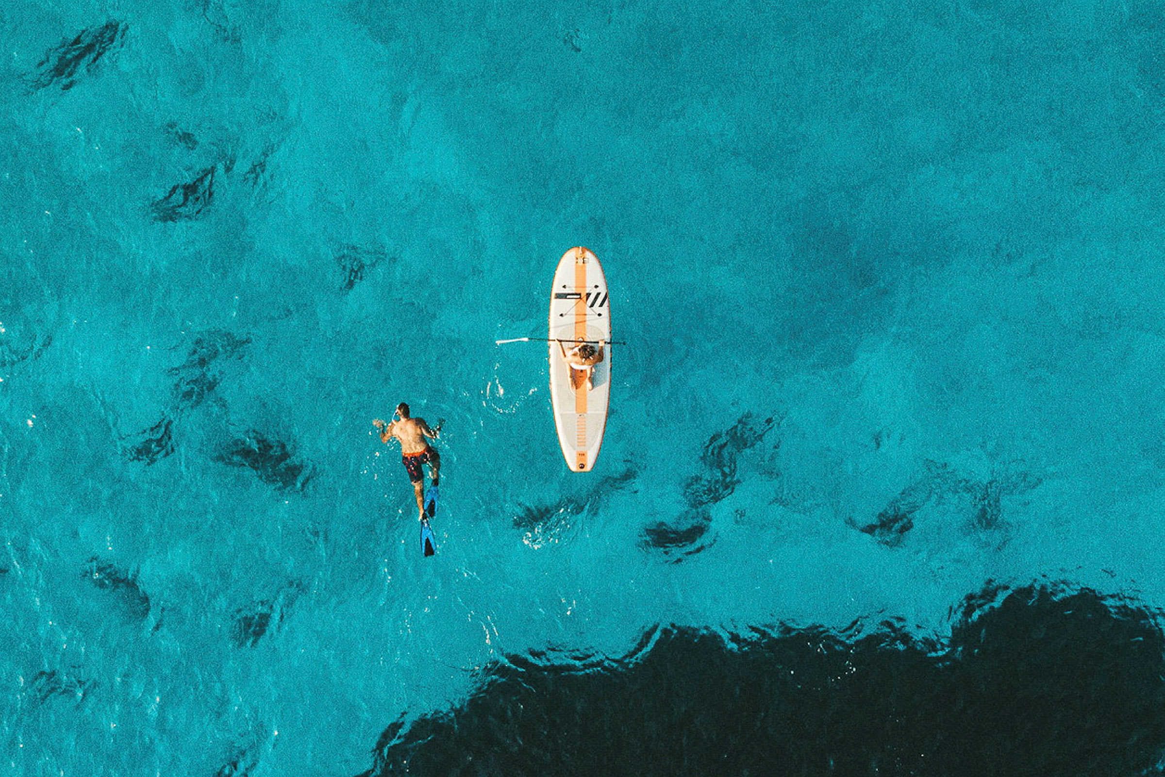 En person padler på et paddleboard, mens en anden snorkler ved siden af. De er i krystalklart turkis vand. (AI-genereret tekst)