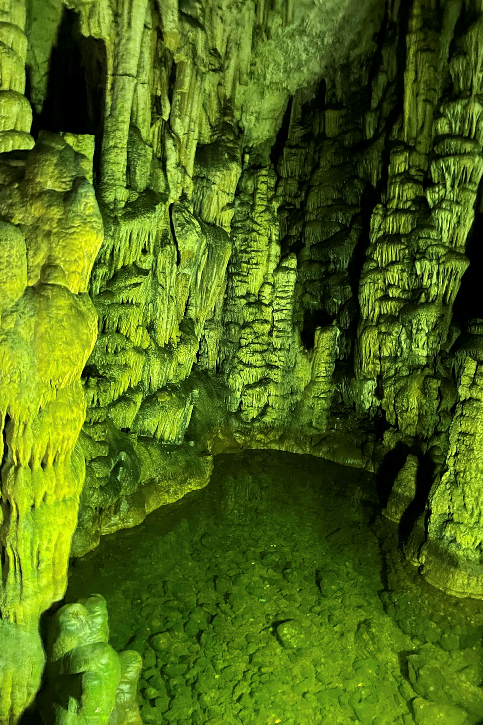En grotte med stalaktitter og stalagmitter, grønt vand og belyste stenformationer. (AI-genereret tekst)