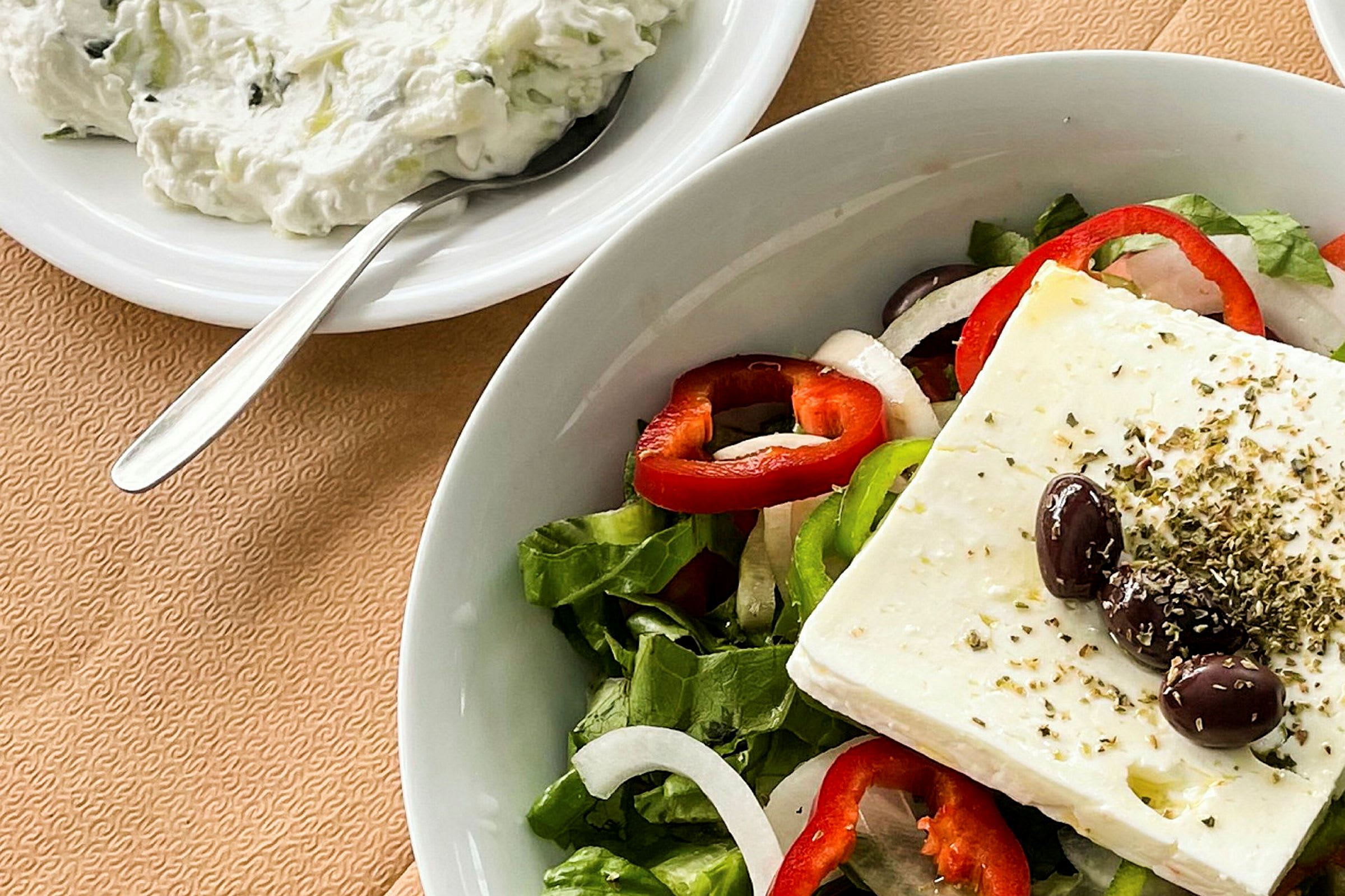 En tallerken med en salat bestående af fetaost, oliven, peberringer, løg og salatblade. Ved siden står en skål med tzatziki og en ske. (AI-genereret tekst)