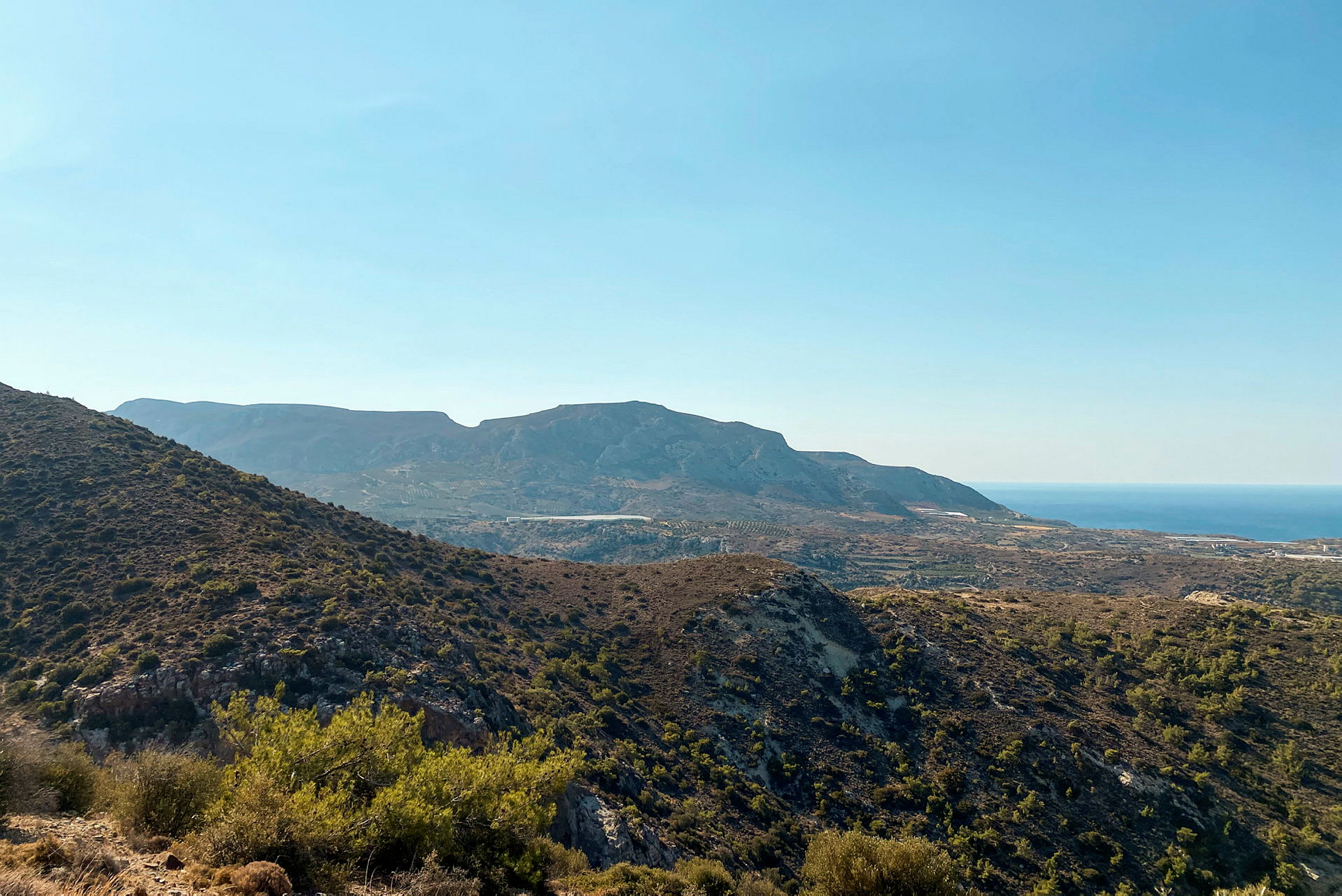 En udsigt over et bjergrigt landskab med varieret vegetation og havet i baggrunden under en klar blå himmel. (AI-genereret tekst)