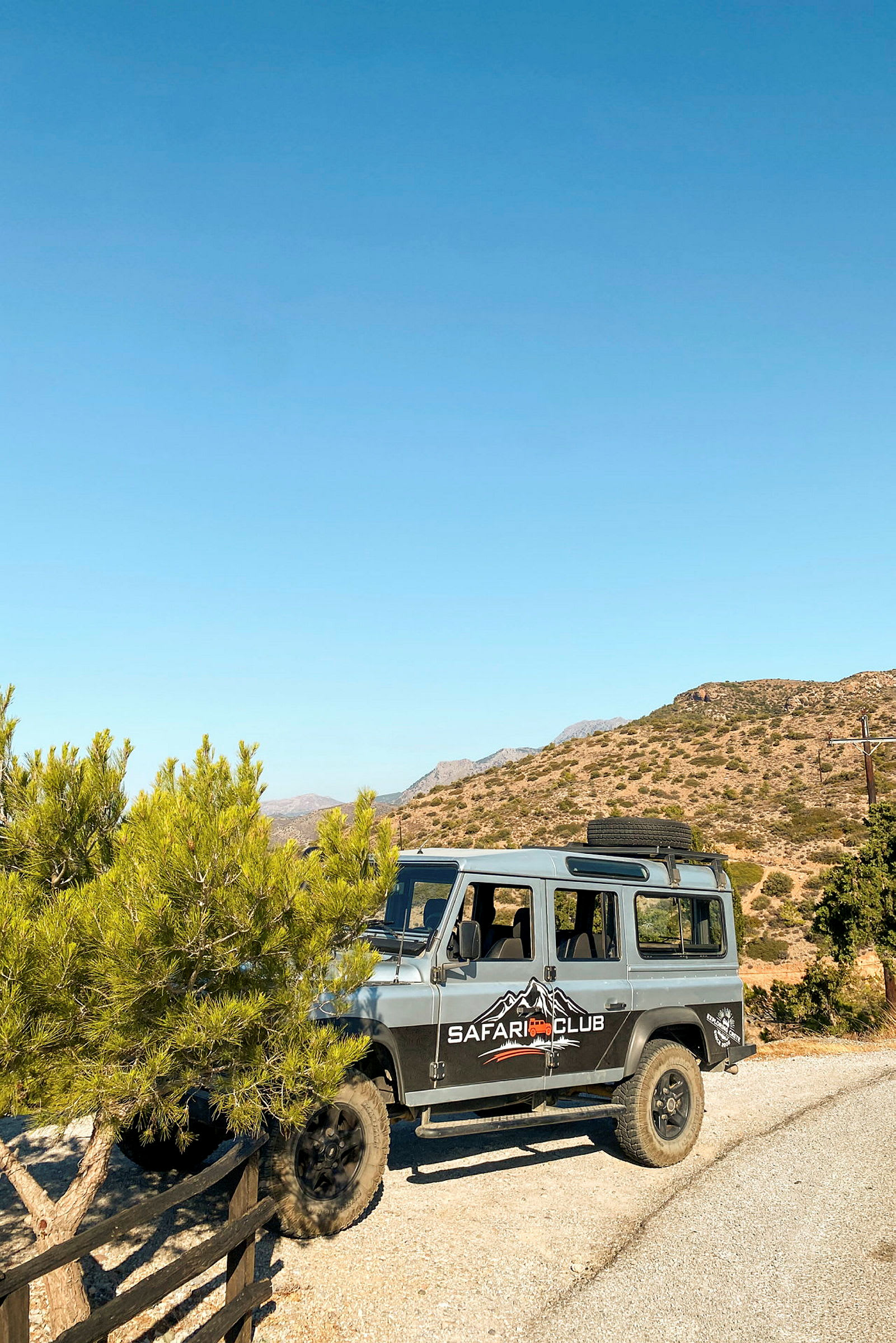 En firehjulstrukken jeep med teksten "Safari Club" på siden holder parkeret på en grusvej ved et træ, med bjerge og en klar himmel i baggrunden. (AI-genereret tekst)