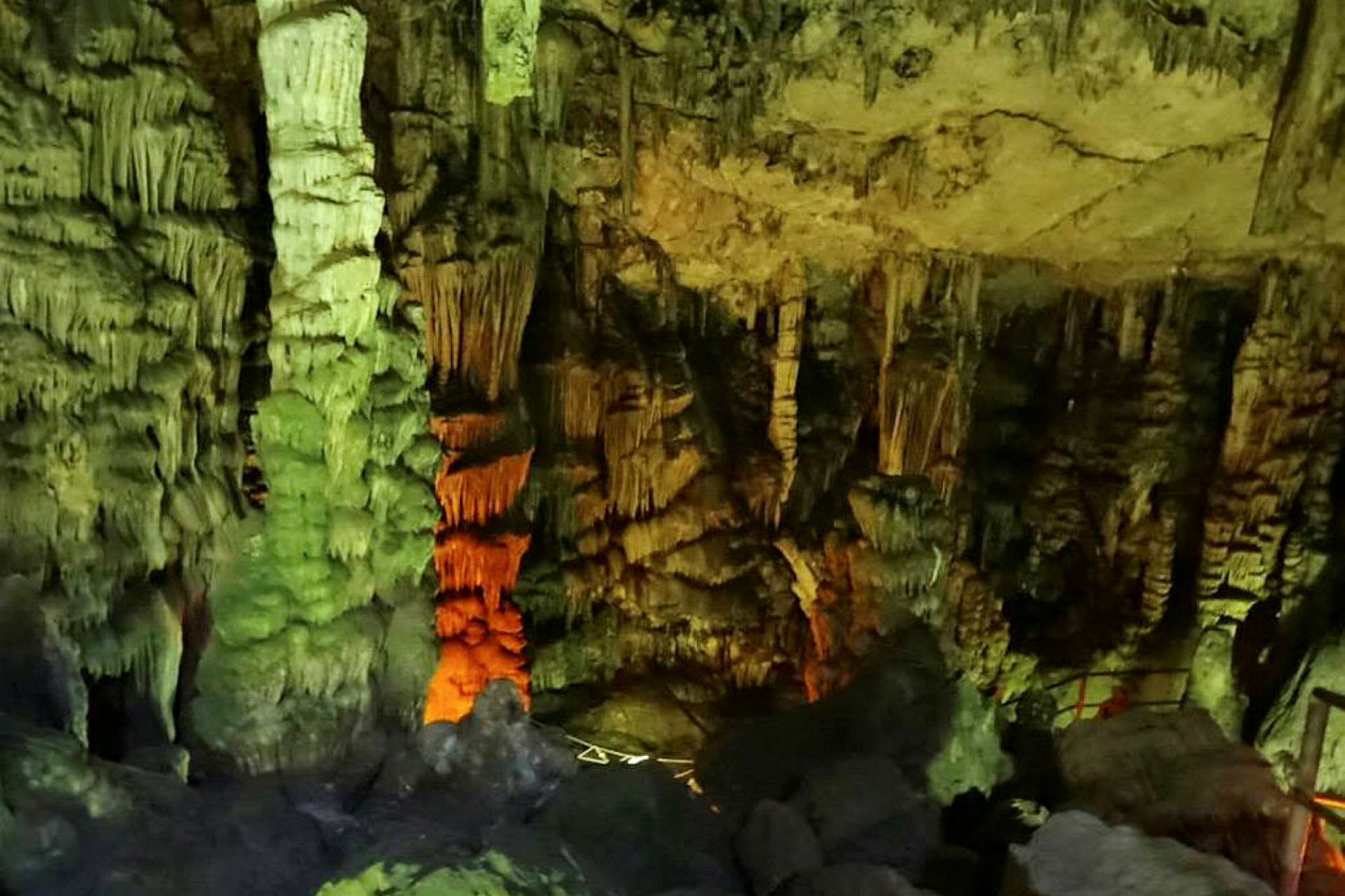 Billede af en grotte med stalaktitter og stalagmitter, oplyst med forskellige farver. (AI-genereret tekst)