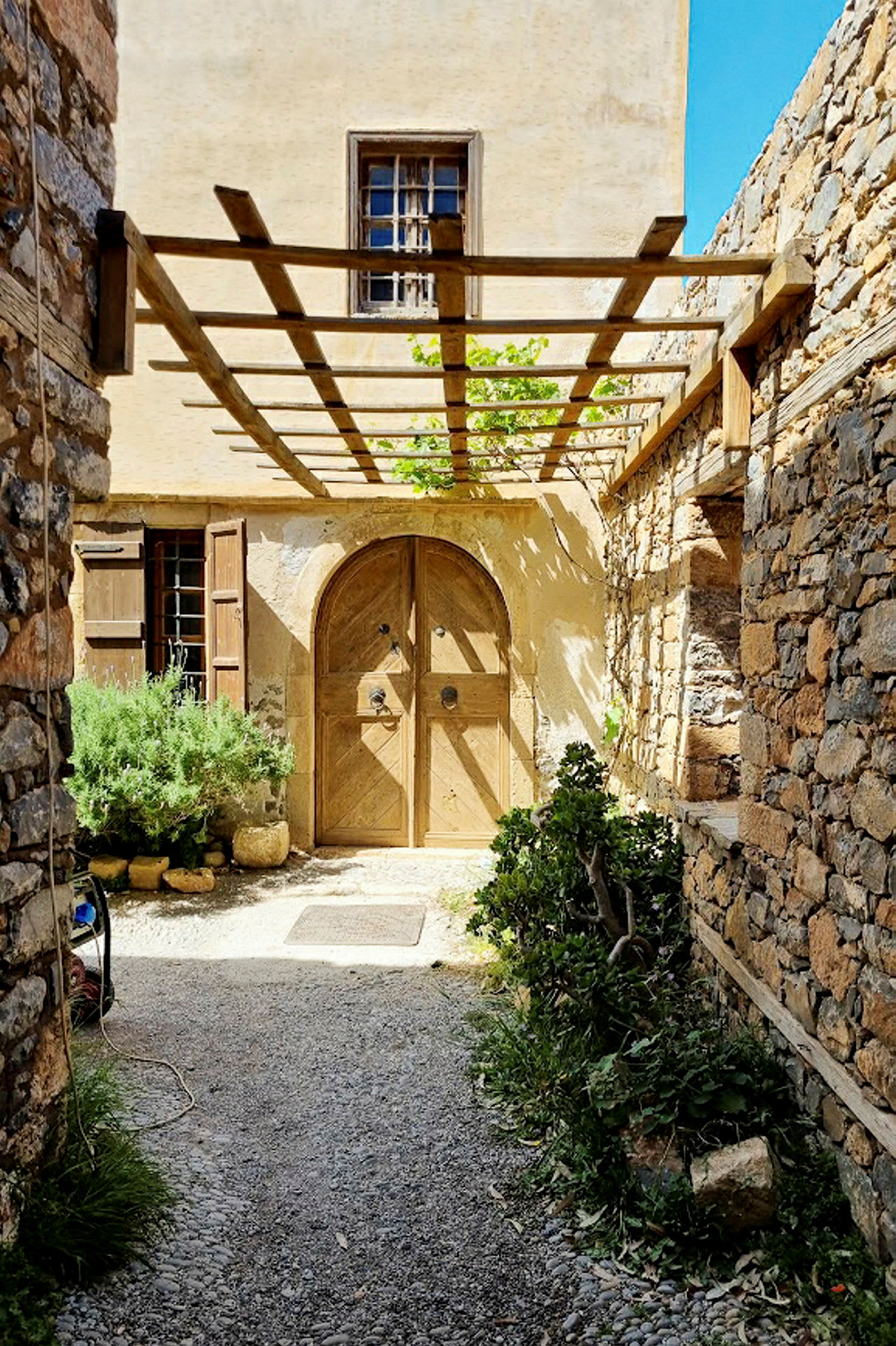 En gammel bygning med en trædør og stenmur omgivet af planter under en pergola. (AI-genereret tekst)