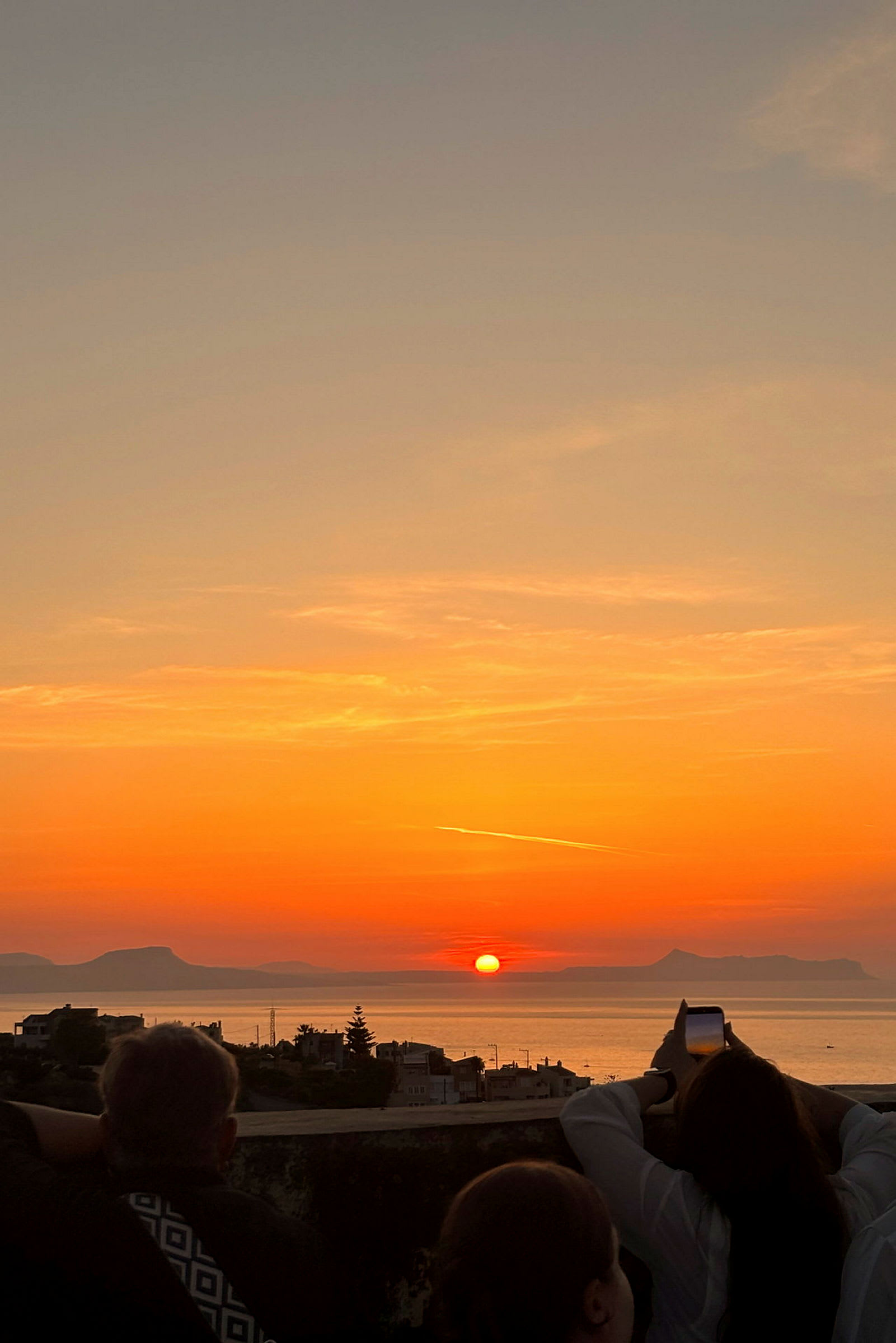 En solnedgang over havet, med silhuetter af bygninger og personer, der observerer og fotograferer udsigten. (AI-genereret tekst)