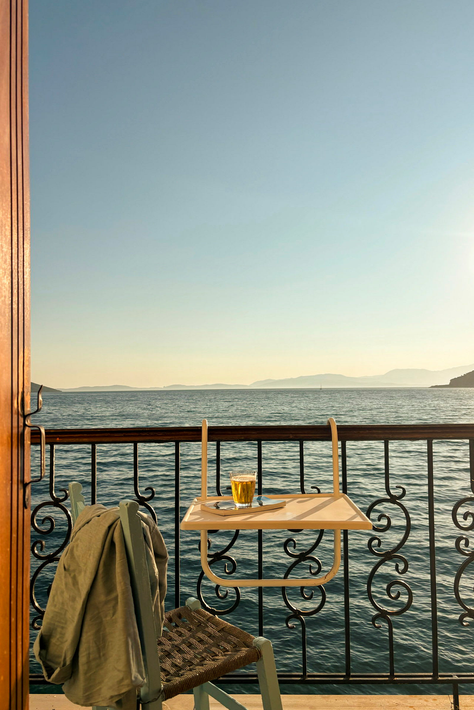 Billedet viser en balkon med udsigt over havet. En stol med en sweater hængende over ryglænet og et lille bord med et glas står ved gelænderet. (AI-genereret tekst)