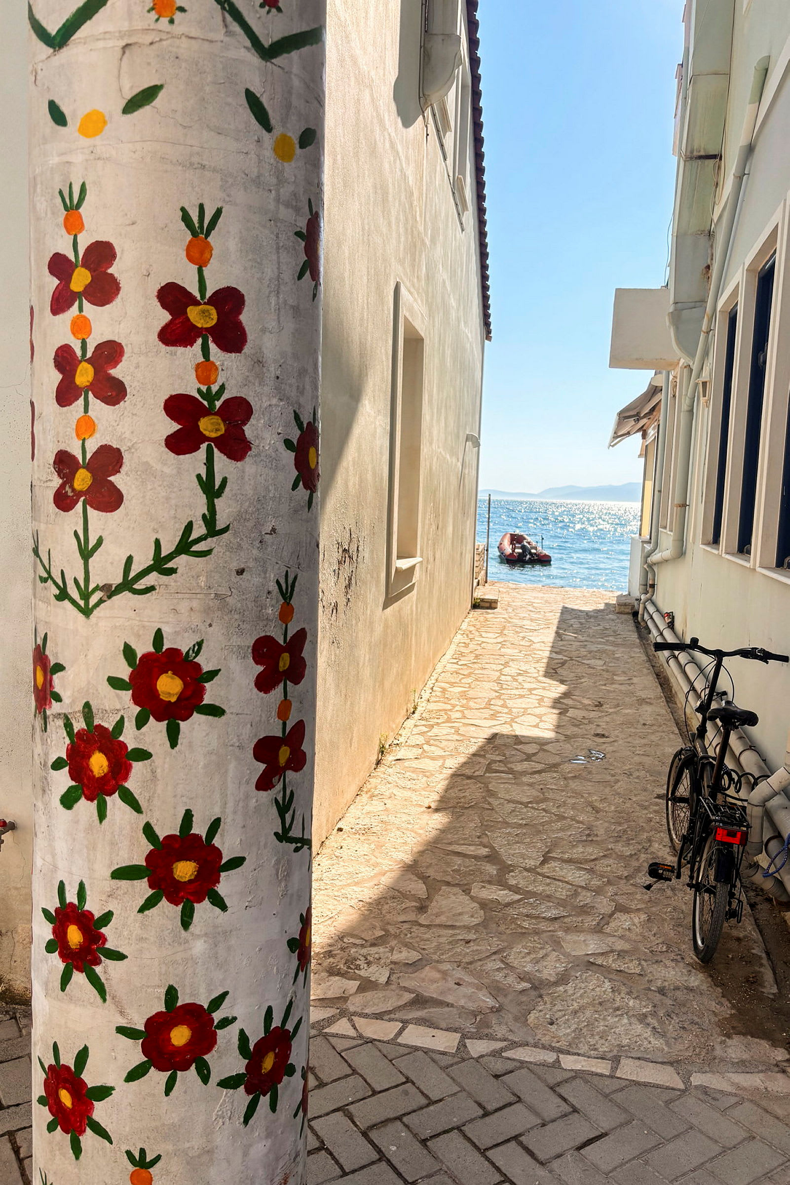 En smal stenbelagt passage mellem huse, med en søjle dekoreret med malede røde blomster i forgrunden og havet synligt i baggrunden. (AI-genereret tekst)