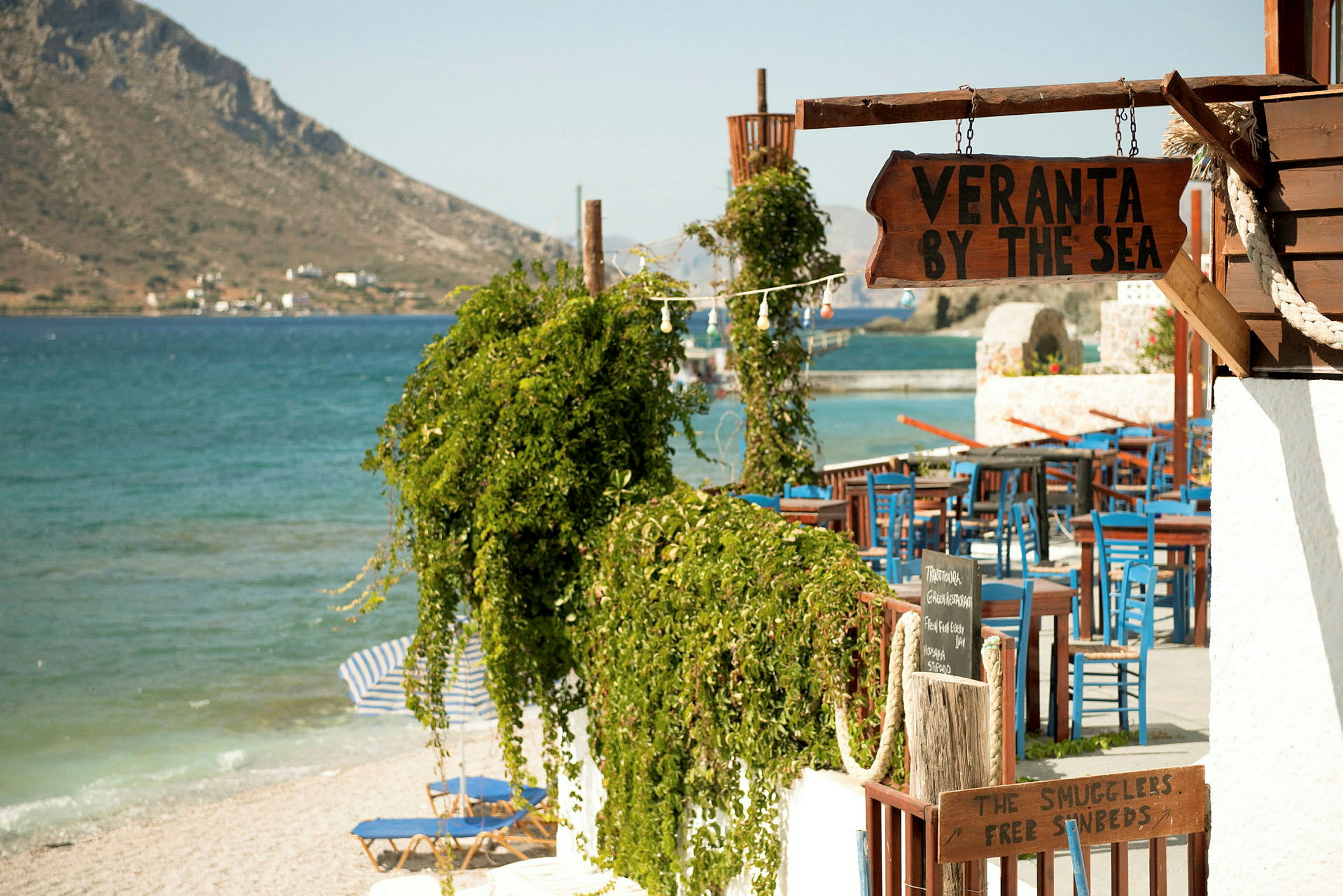 Et nærbillede af en restaurant eller veranda ved stranden med skilte der viser "Veranta by the sea" og blå stole samt et grønt område i forgrunden. (AI-genereret tekst)