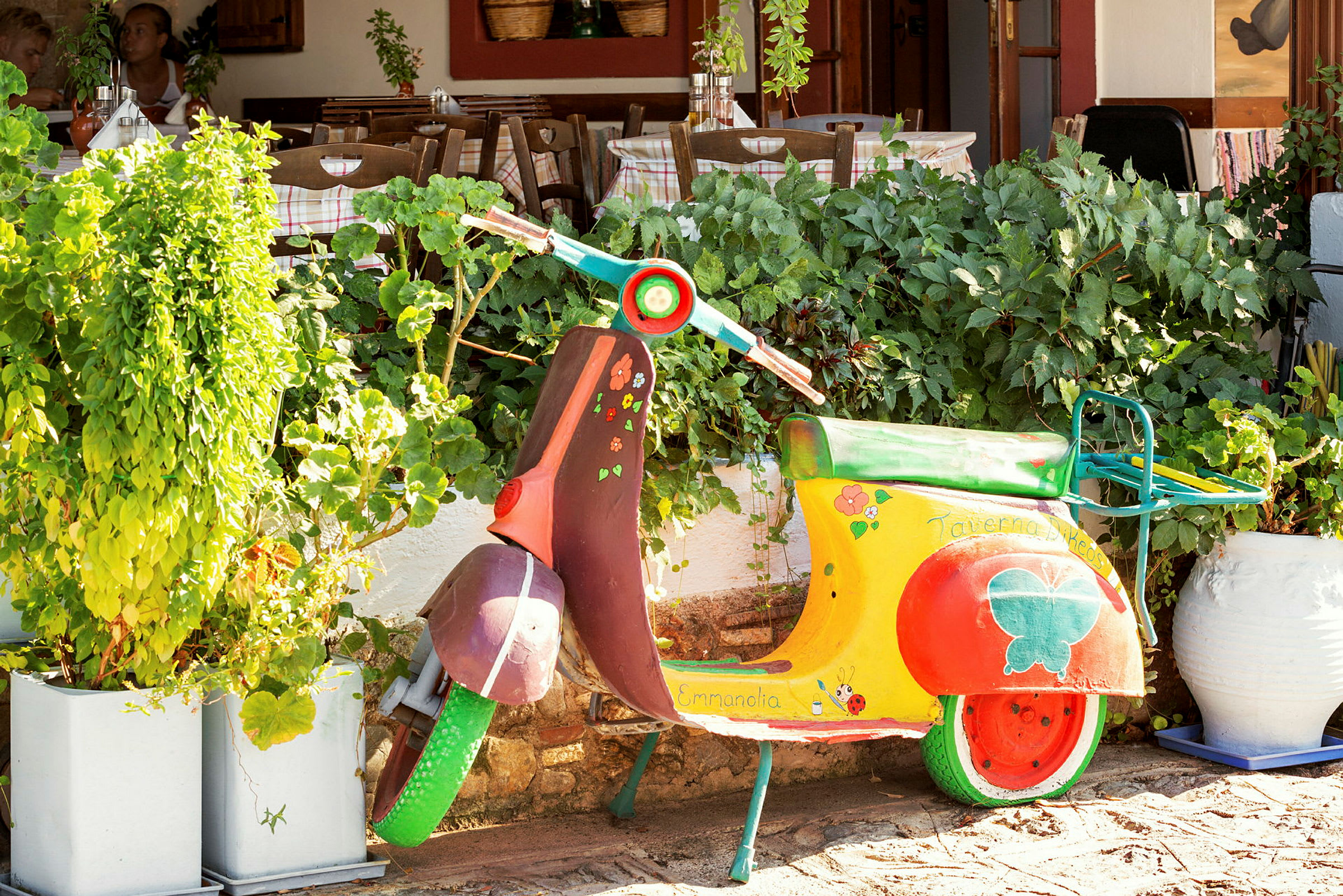 En farverig vespa placeret foran grønne planter og en restaurants siddeområde. (AI-genereret tekst)
