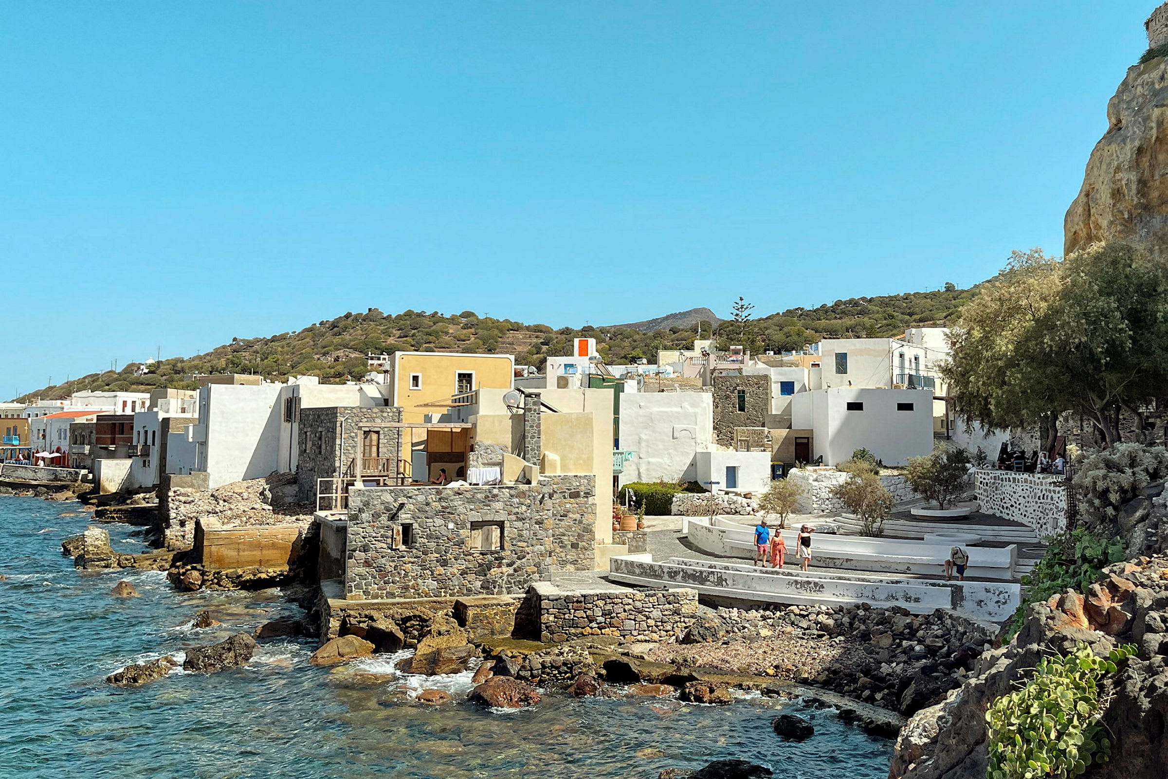 En kystlinje med huse i forskellige farver og stenudformninger, ved siden af klart hav og omgivet af en bakke. (AI-genereret tekst)