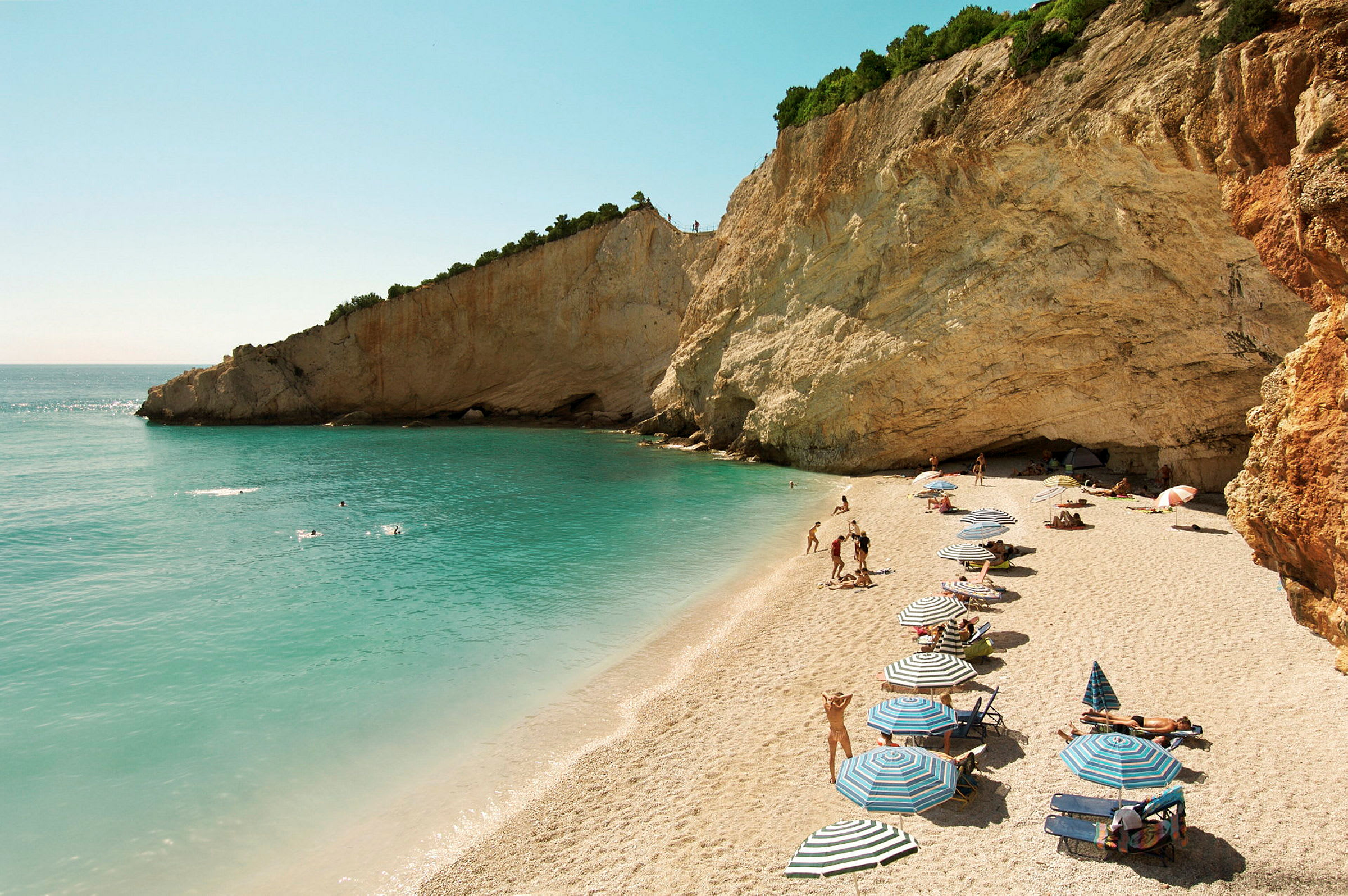 Strand med parasoller, sand, klippevæg og svømmende mennesker ved en turkis hav. (AI-genereret tekst)