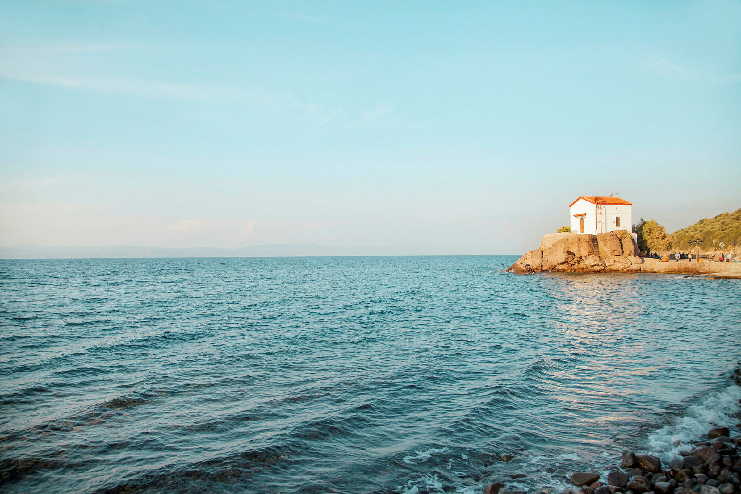 Et lille hvidt hus med rødt tag ligger på en klippe ved havet under en klar blå himmel. (AI-genereret tekst)