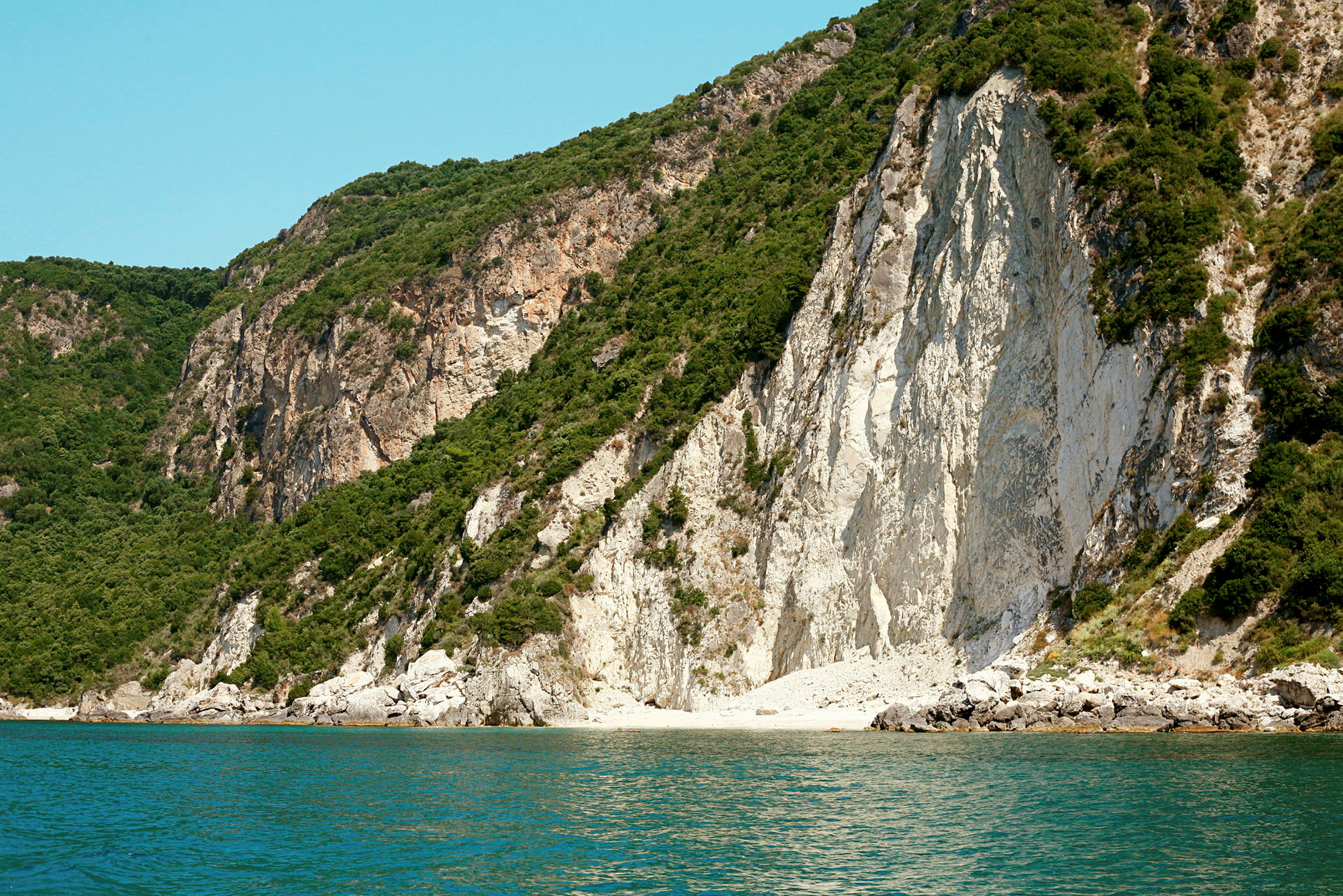 En stejl klippe med grøn bevoksning ved havet, med klart blåt vand i forgrunden. (AI-genereret tekst)