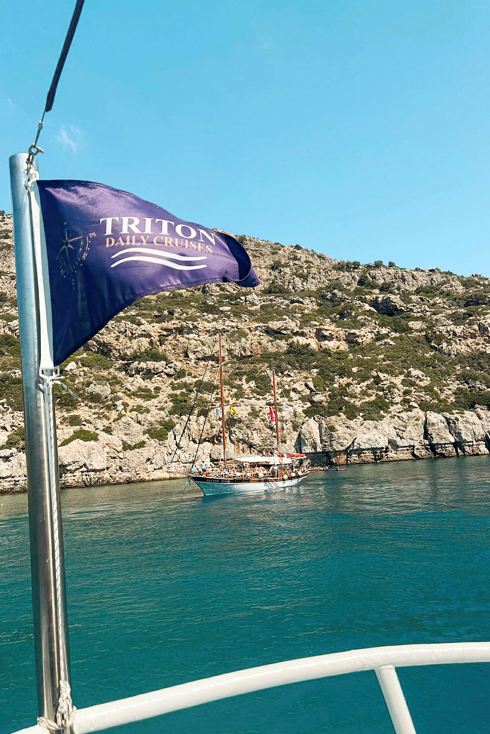 Et sejlskib ligger ved en klippe, mens et flag med teksten "Triton Daily Cruises" vajer i forgrunden. (AI-genereret tekst)