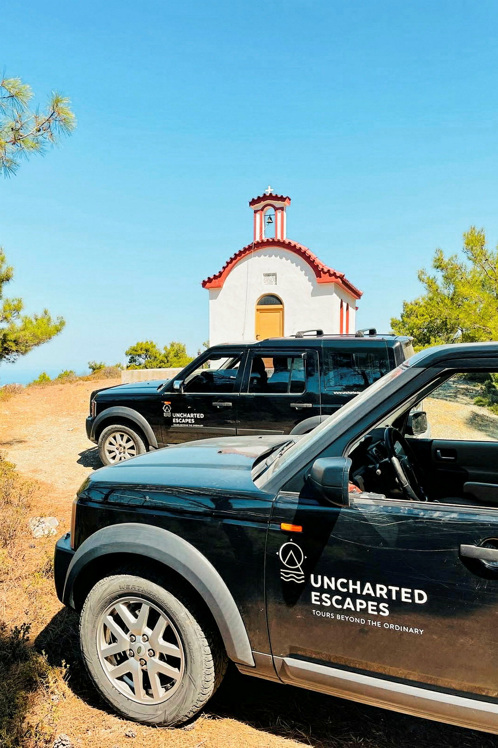 To sorte biler med logoen "Uncharted Escapes" er parkeret foran en lille, hvid kirke med rødt tag. Kirken er omgivet af grønt, og himlen er klar og blå. (AI-genereret tekst)