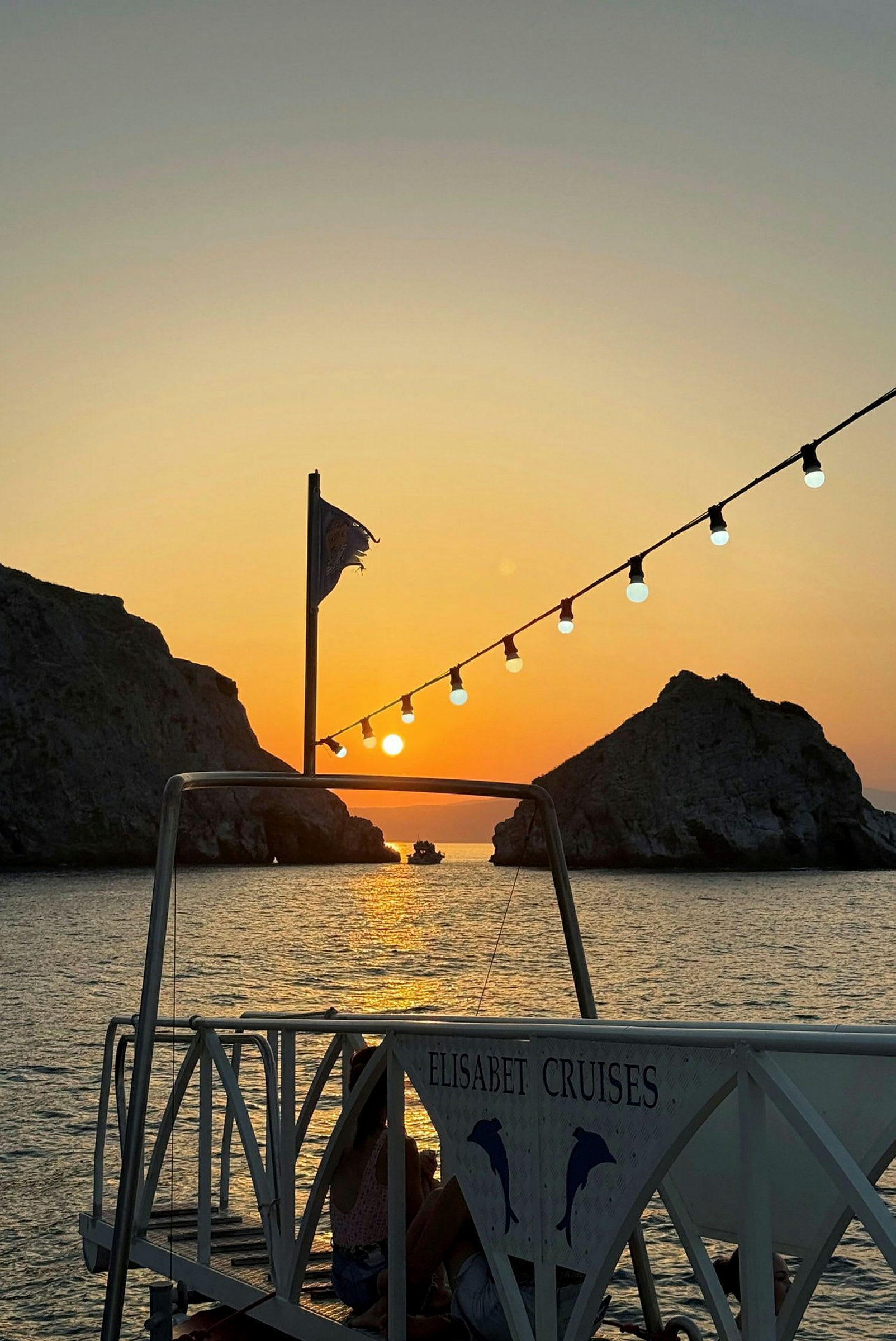 En solnedgang over havet med en bro i forgrunden, der bærer et skilt med teksten 'Elisabet Cruises'. I baggrunden ses klipper og en båd. (AI-genereret tekst)