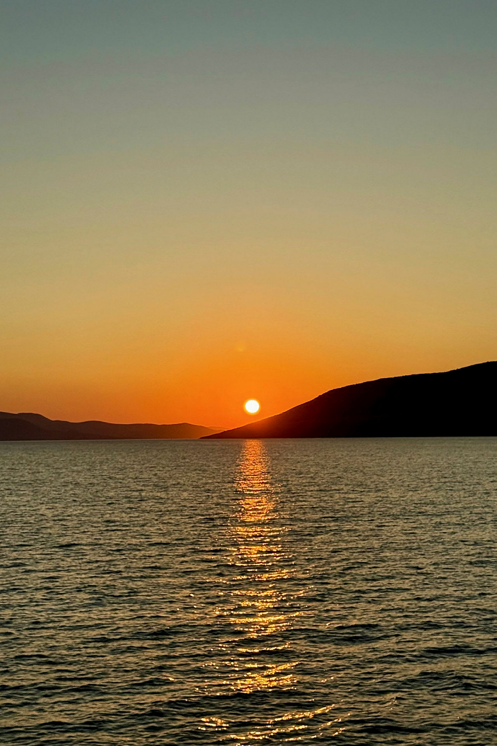 Billede af en solnedgang over havet med refleksioner i vandet og bjerge i baggrunden. (AI-genereret tekst)