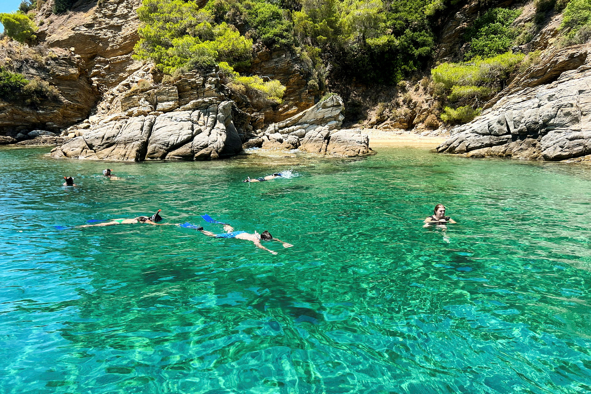 Personer svømmer og snorkler i klart turkis vand nær en klippefyldt strand. (AI-genereret tekst)