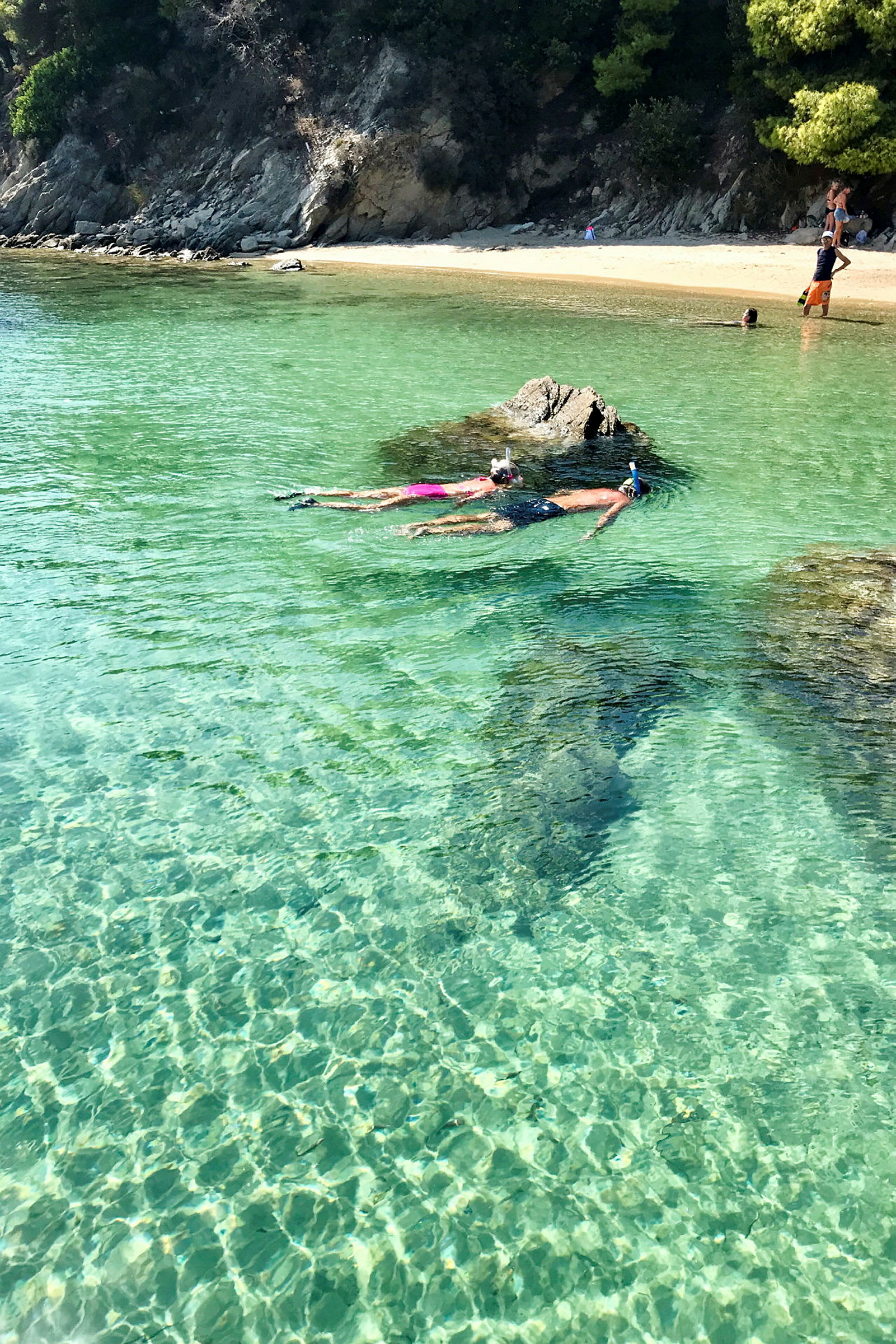 To personer snorkler i klart, turkis vand nær en sten, med en sandstrand og træer i baggrunden. (AI-genereret tekst)