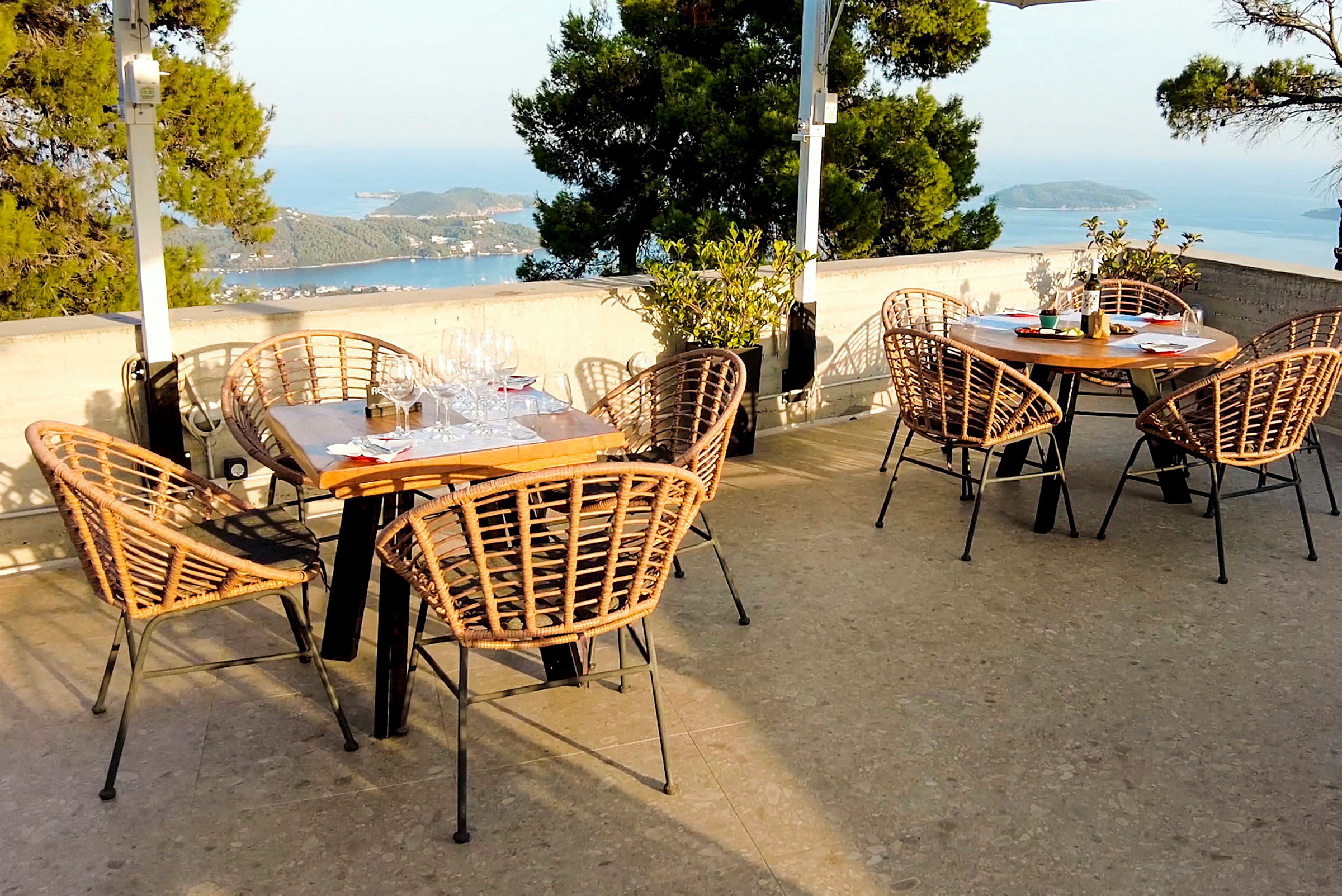 Udendørs restaurant med borde og flettede stole, med udsigt over havet og grønne omgivelser. (AI-genereret tekst)
