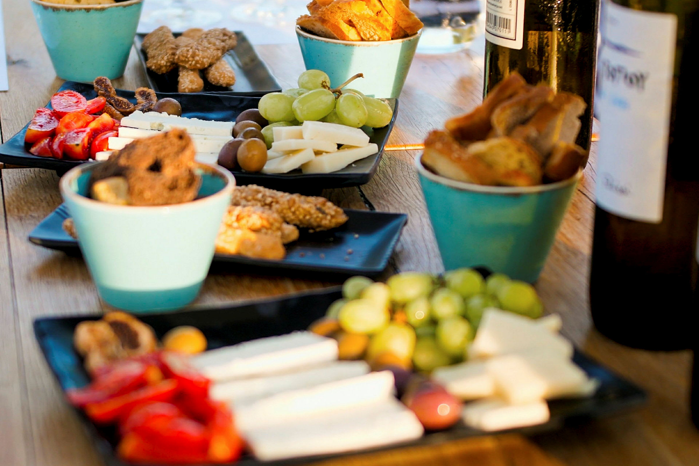 Flere tallerkener indeholder snacks som ost, grøntsager, oliven og brød, sammen med drikkeflasker på et bord. (AI-genereret tekst)
