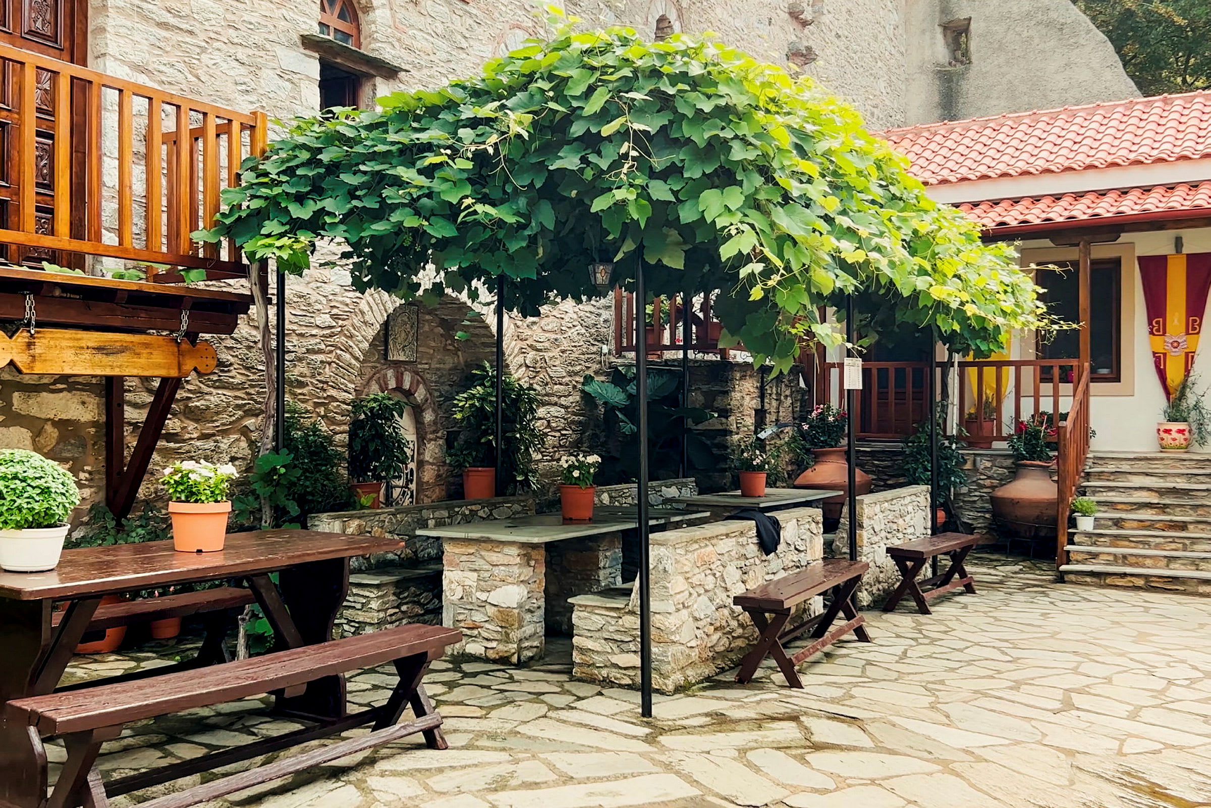 En gårdhave med stenbelagt gulv, træborde og bænke samt store blomsterkrukker. Grønne planter klatrer på en pergola ovenover. (AI-genereret tekst)
