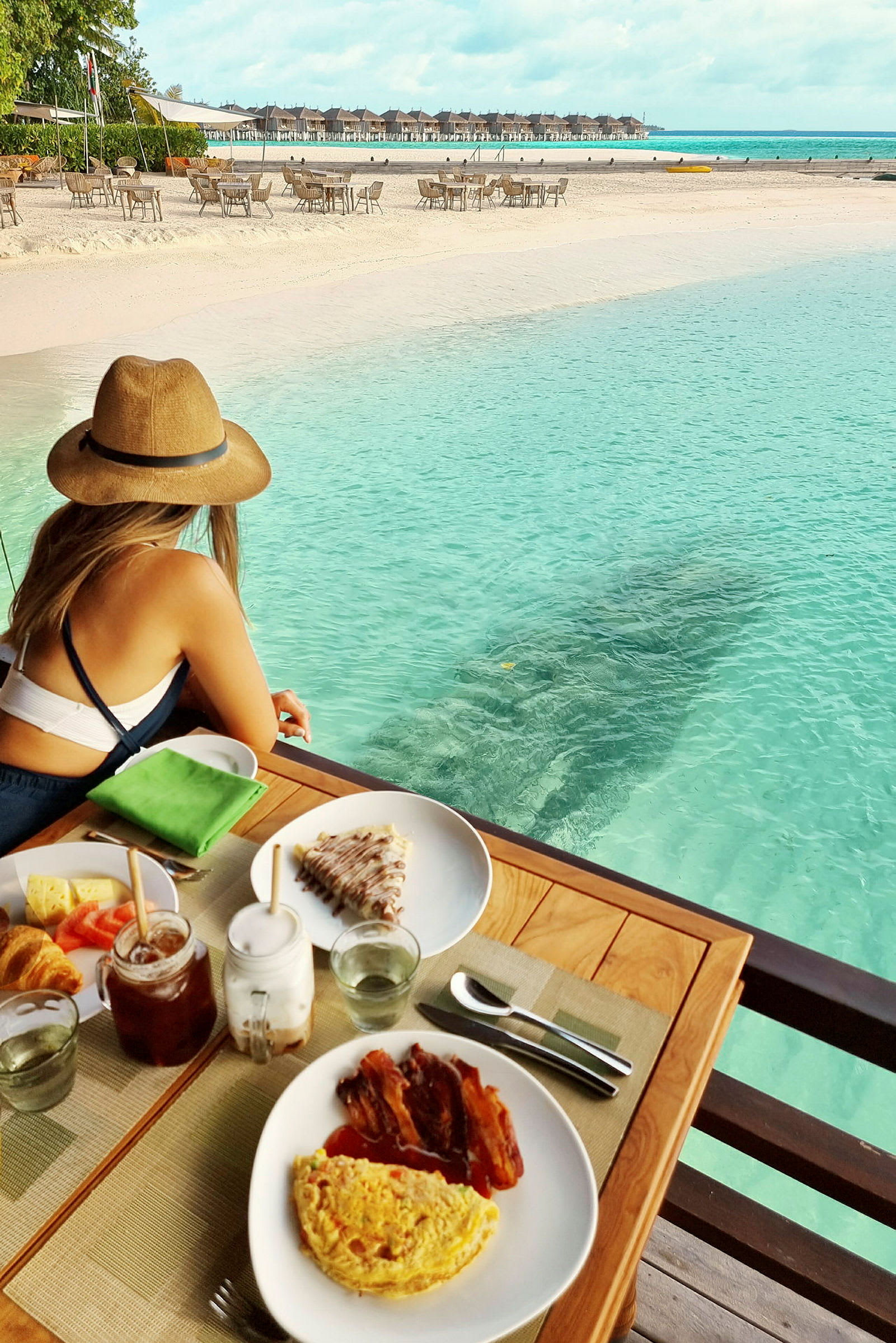 En kvinde sidder ved et bord med morgenmad ved strandkanten med turkis vand og udsigt over stranden og bungalower. (AI-genereret tekst)