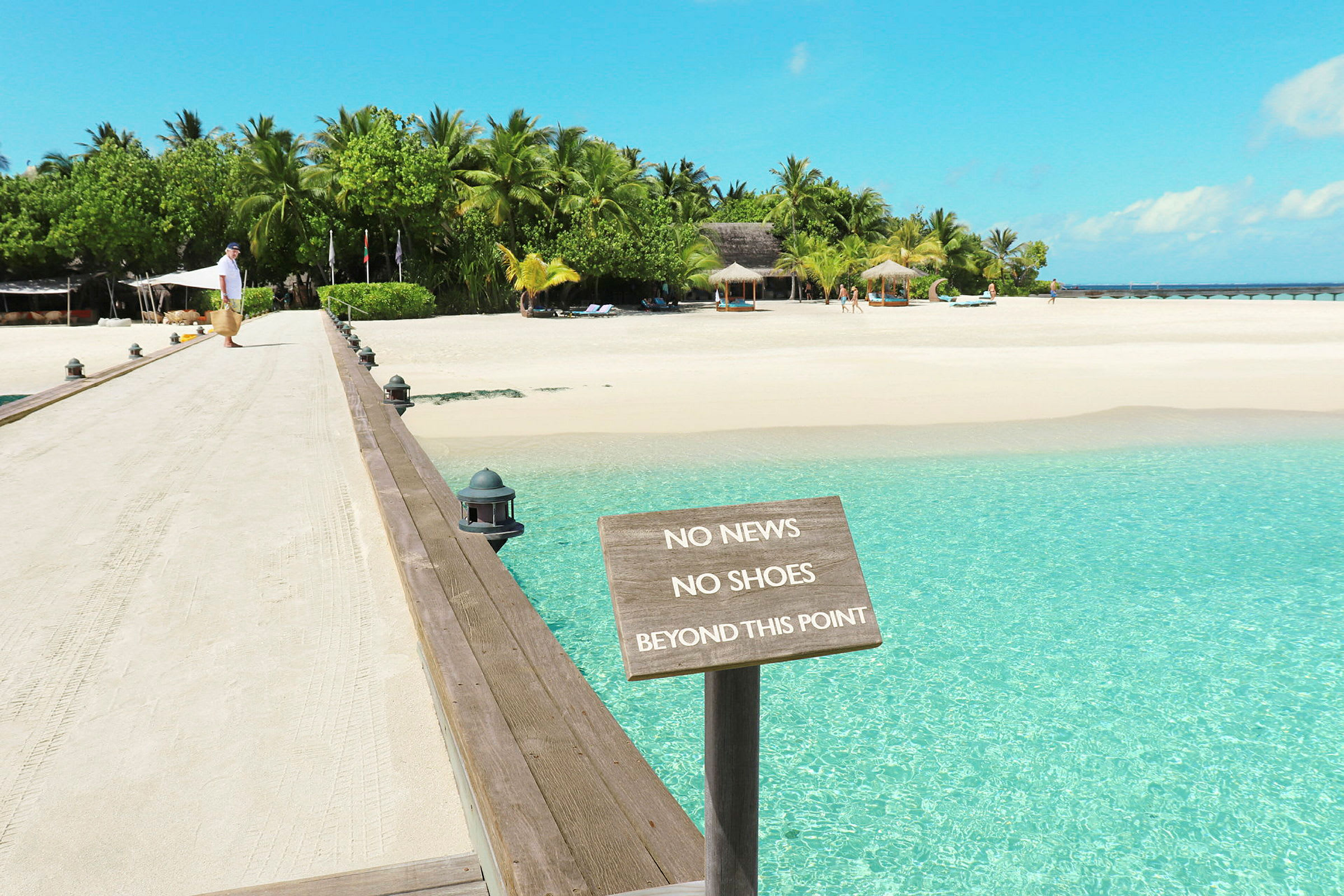 En sandet gangbro fører til en tropisk strand med et skilt, der lyder 'No News No Shoes Beyond This Point'. (AI-genereret tekst)