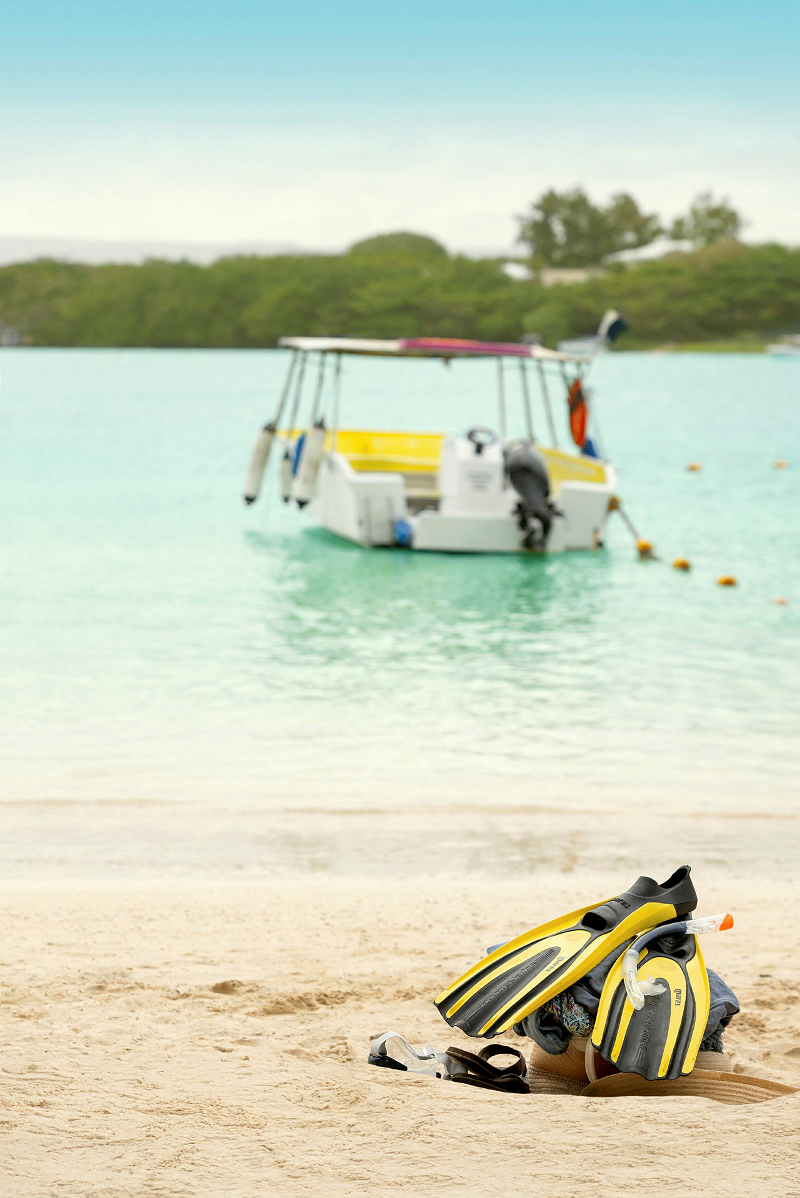 Et par gul-sorte svømmefødder, snorkeludstyr og sandaler er placeret på en sandstrand. En båd er forankret i vandet i baggrunden. (AI-genereret tekst)