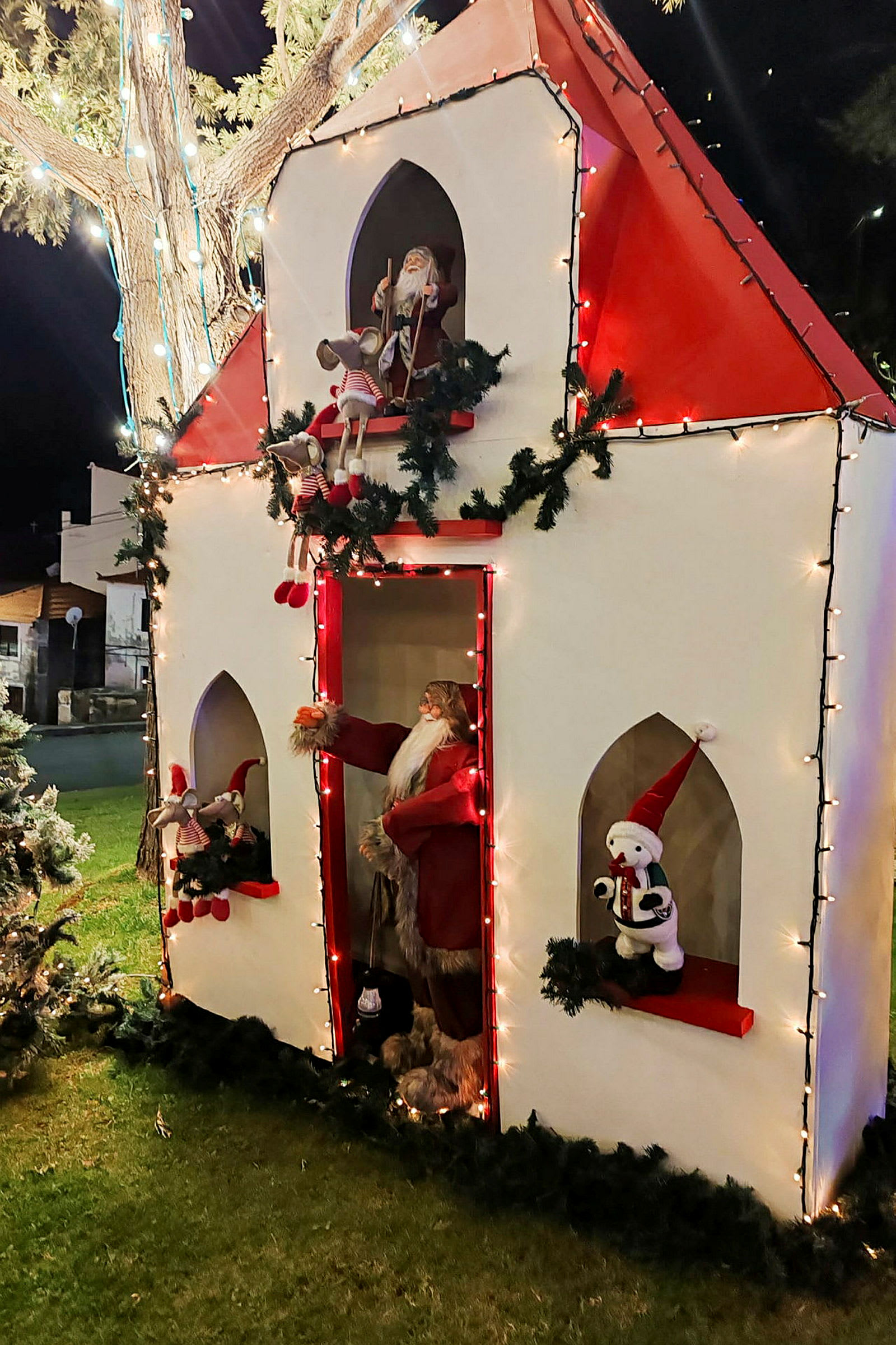 En juledekoration, der forestiller et hus med julemanden og legetøj, dekoreret med lyskæder og kranse. (AI-genereret tekst)