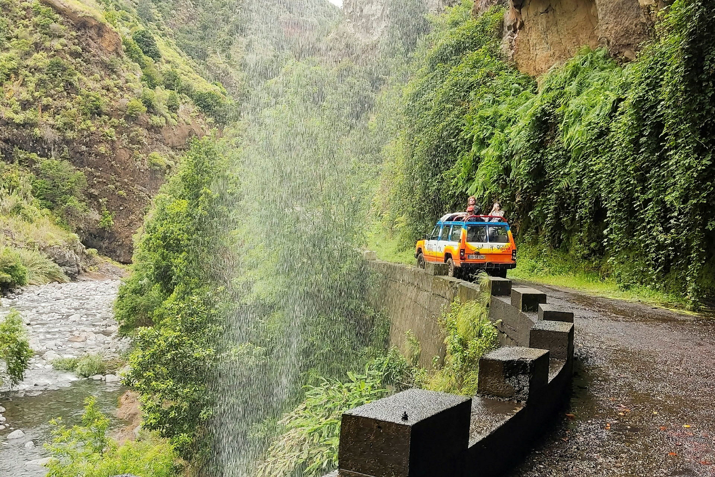 En orange jeep parkeret på en smal vej ved siden af en grøn dal med regn eller vandsprøjt. (AI-genereret tekst)