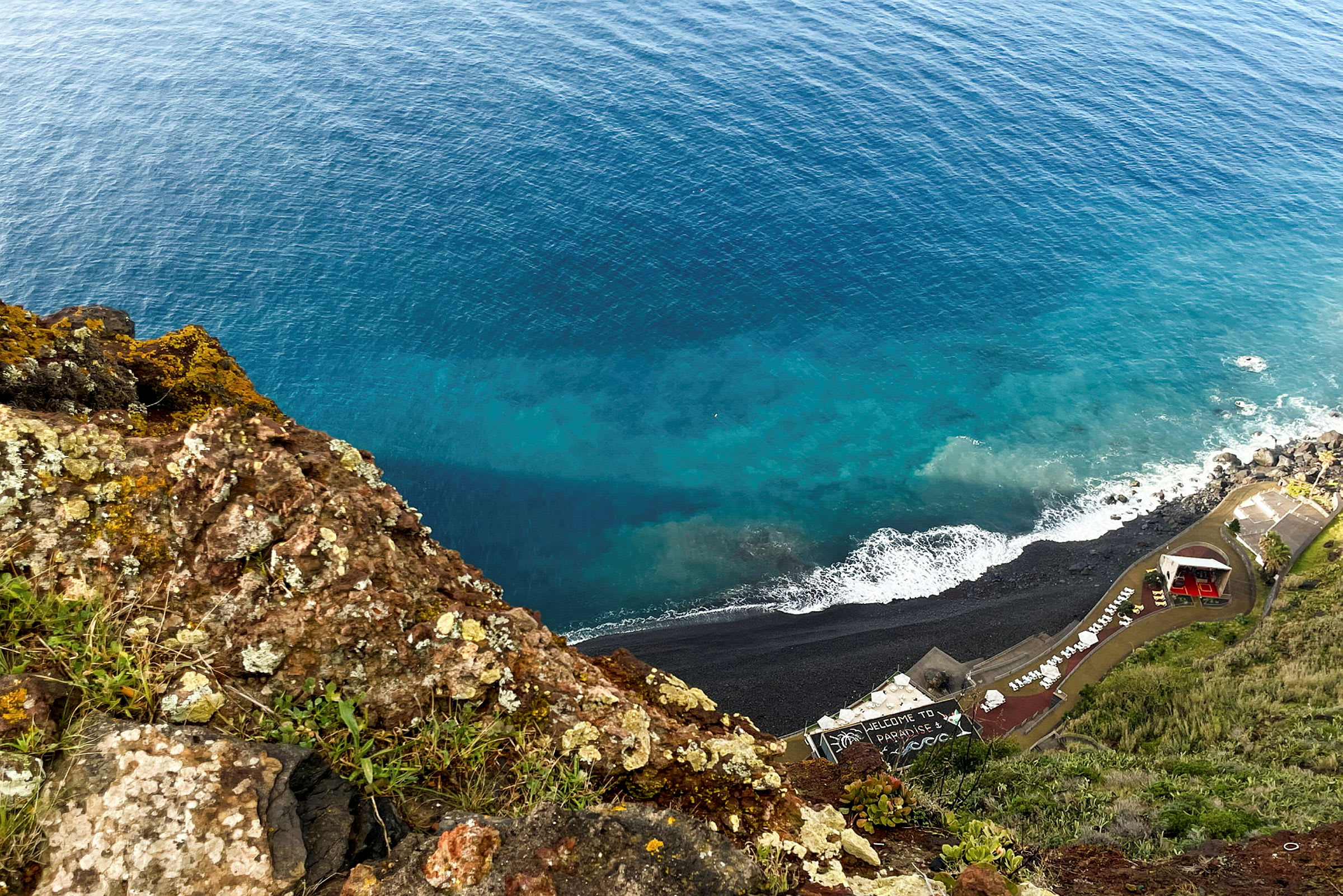Udsigt fra en klippe ned mod en kystlinje med stenstrand og bygninger, samt havet. (AI-genereret tekst)