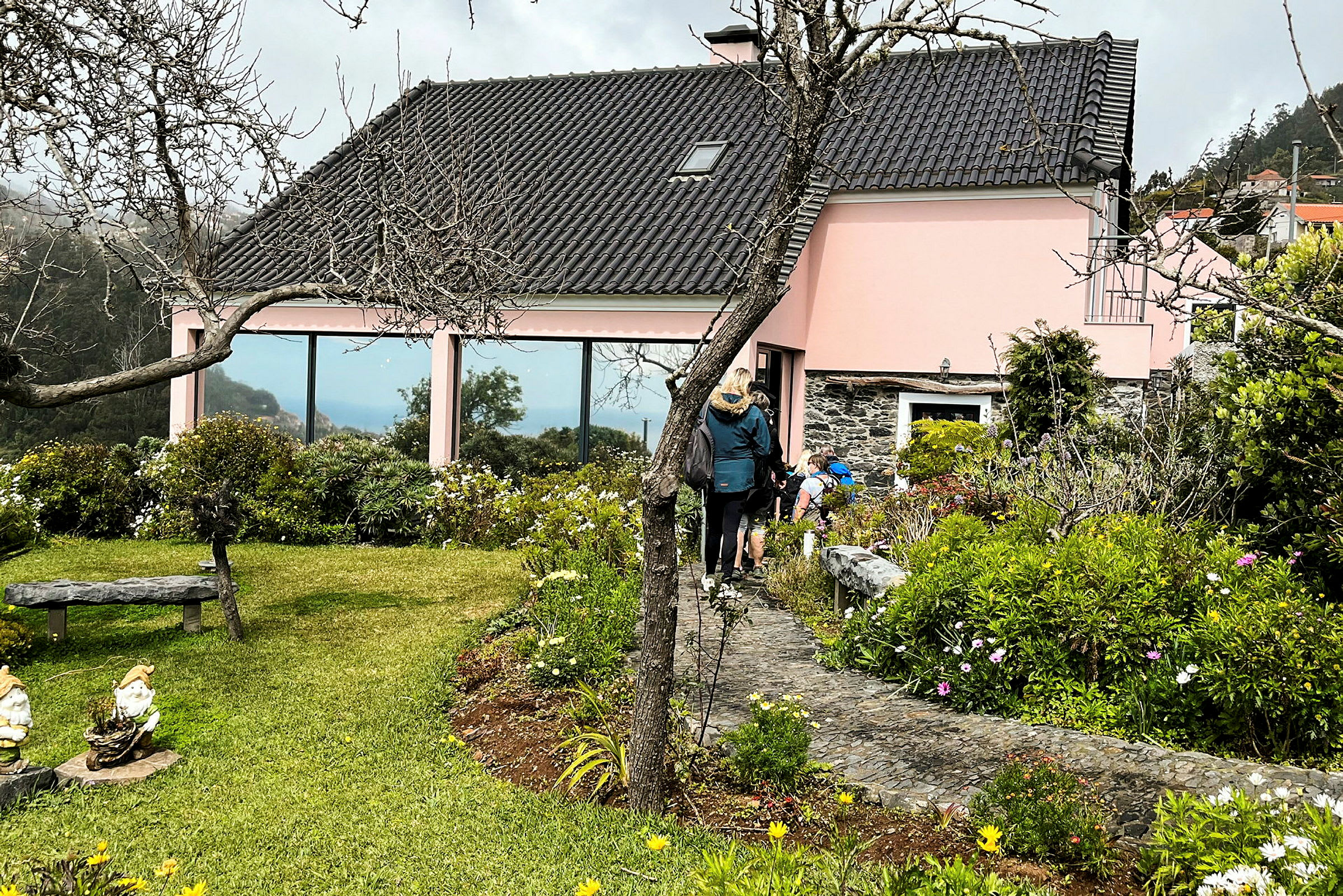 En lyserød bygning med sort tag omgivet af grønne planter og blomster, med personer gående på en sti. (AI-genereret tekst)