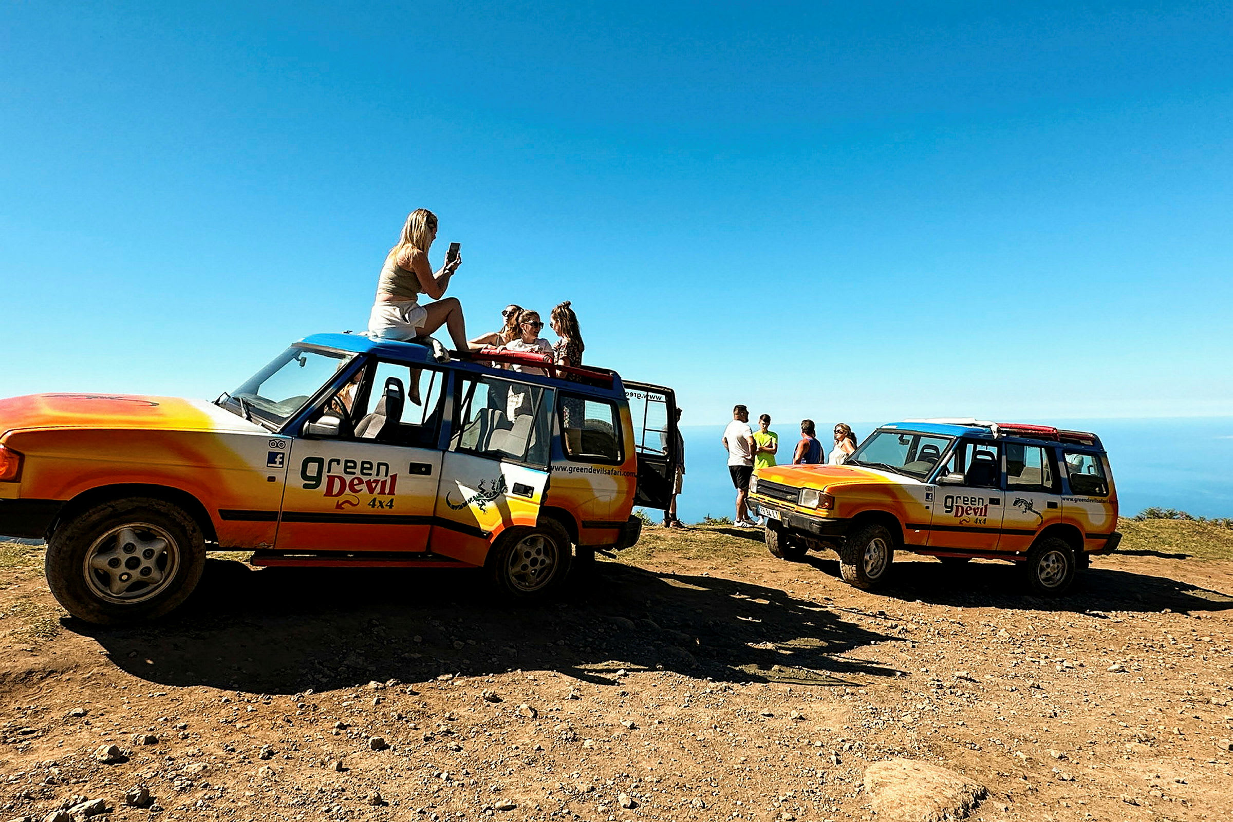 To terrængkøretøjer med "Green Devil 4x4" logoer er parkeret på en grusvej med udsigt over havet. Folk står eller sidder i nærheden af køretøjerne. (AI-genereret tekst)